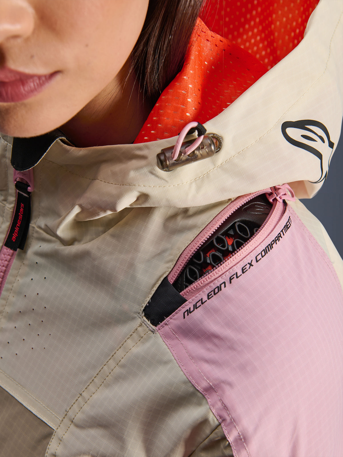 Pormenor do casaco Alpinestars Molly Air em Sand Pink Almond, destacando o compartimento com fecho de correr no ombro para protetores Nucleon Flex, o forro do capuz em malha cor-de-laranja vibrante e a construção em tecido ripstop durável.