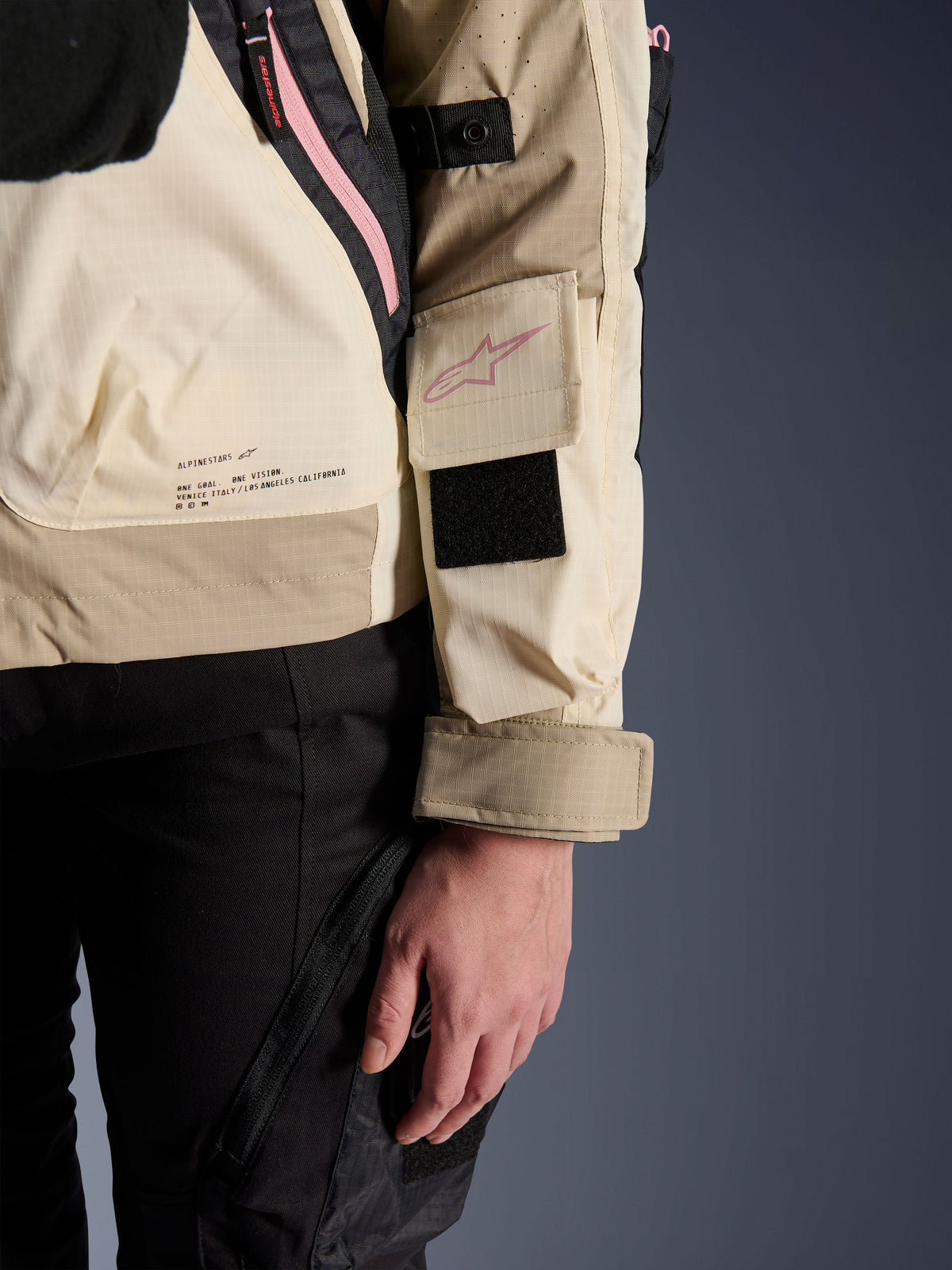 Alpinestars Molly Air Jacket, casaco de motociclismo urbano para mulher, Sand Pink Almond, vista lateral de pormenor que mostra o bolso na manga com logótipo cor-de-rosa, patch de Velcro preto e o texto da missão da marca na parte inferior das costas, material ripstop leve