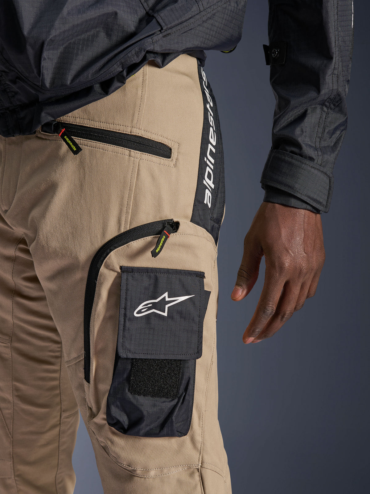 Calças Alpinestars Acteon em Sand, vista detalhada da zona da anca e da coxa, apresentando um bolso cargo lateral preto com logótipo de estrela branco, bolsos com fecho de correr e marca Alpinestars vertical num painel lateral preto, vestuário urbano para motociclismo.