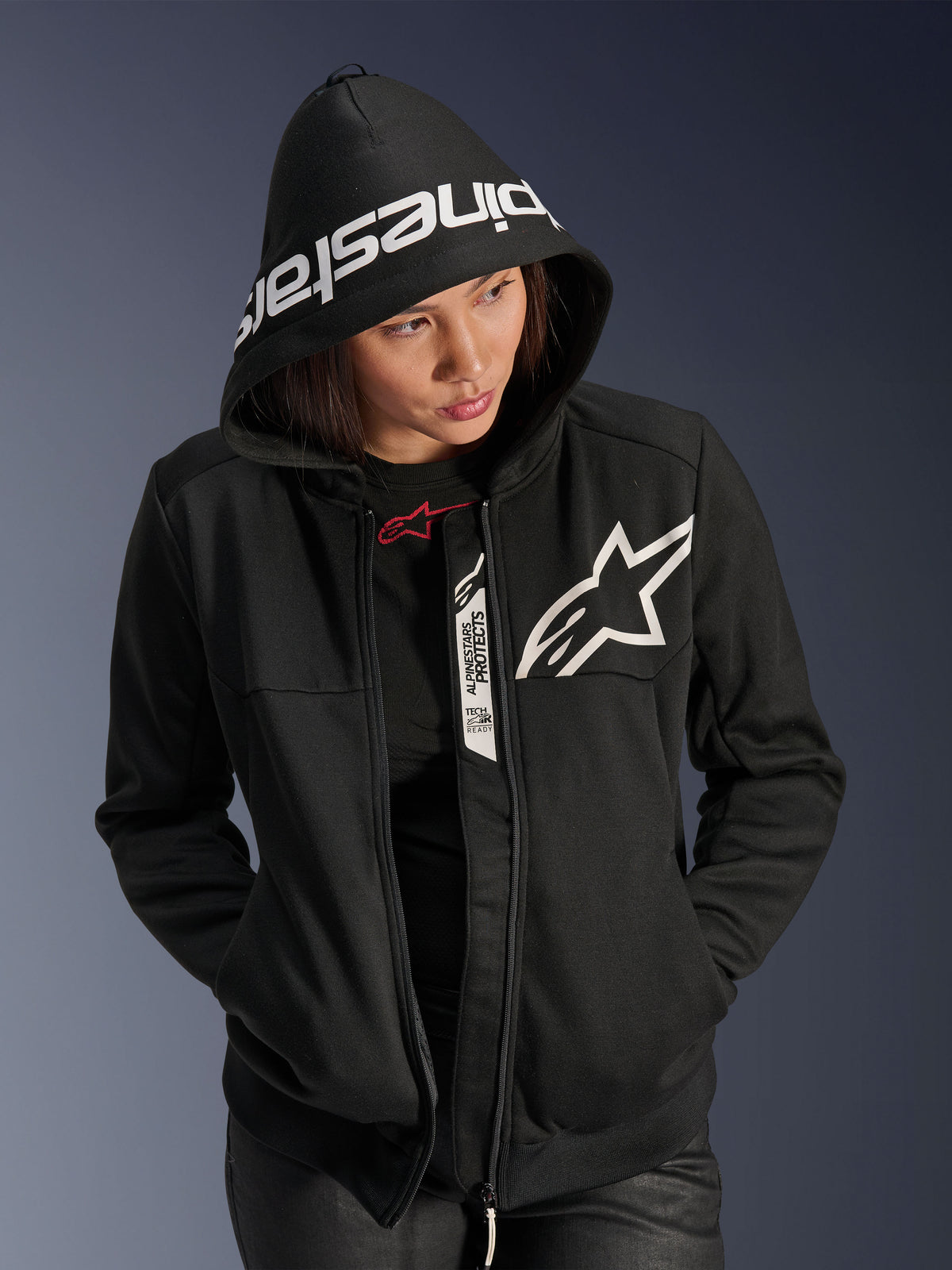 Alpinestars Stella Chrome V2 Sport Hoodie para mulher, preto e branco, hoodie de motociclismo com logótipo de estrela branco no peito e marca Alpinestars na aba do capuz, mulher com o capuz colocado, estilo desportivo urbano casual com detalhes Tech-Air Ready