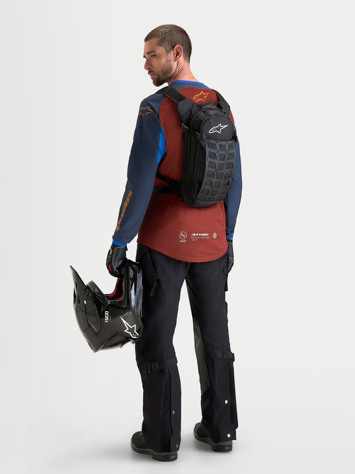 Alpinestars Mochila AT-9, mochila de motocross preta e cinzento-escuro, vista traseira num modelo a usar equipamento de equitação e a segurar um capacete