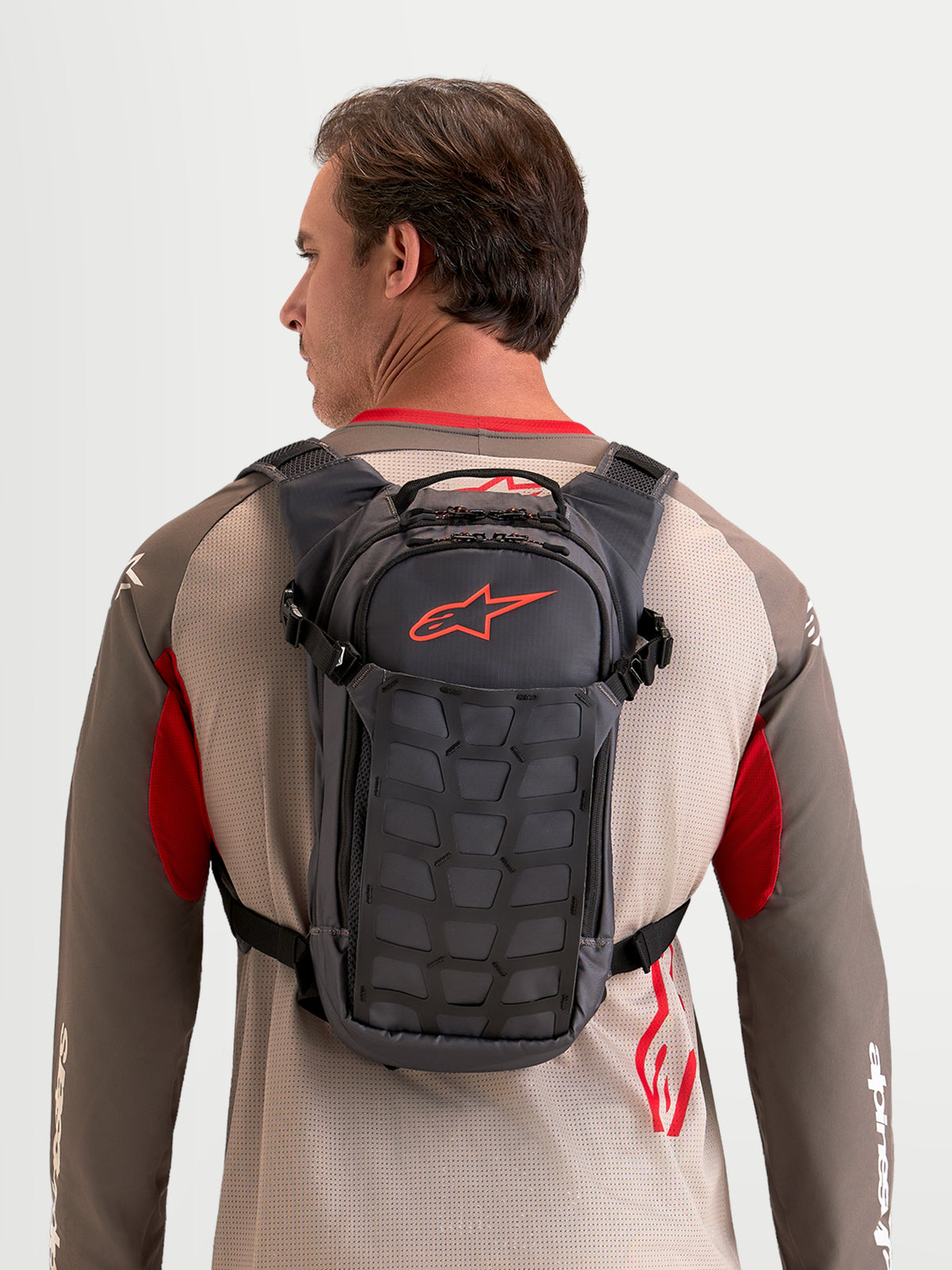 Alpinestars Mochila AT-9, mochila de moto para motocross e uso diário num modelo, construção em tecido cinzento e castanho, com compartimentos de arrumação organizados e um logótipo Astar vermelho distintivo