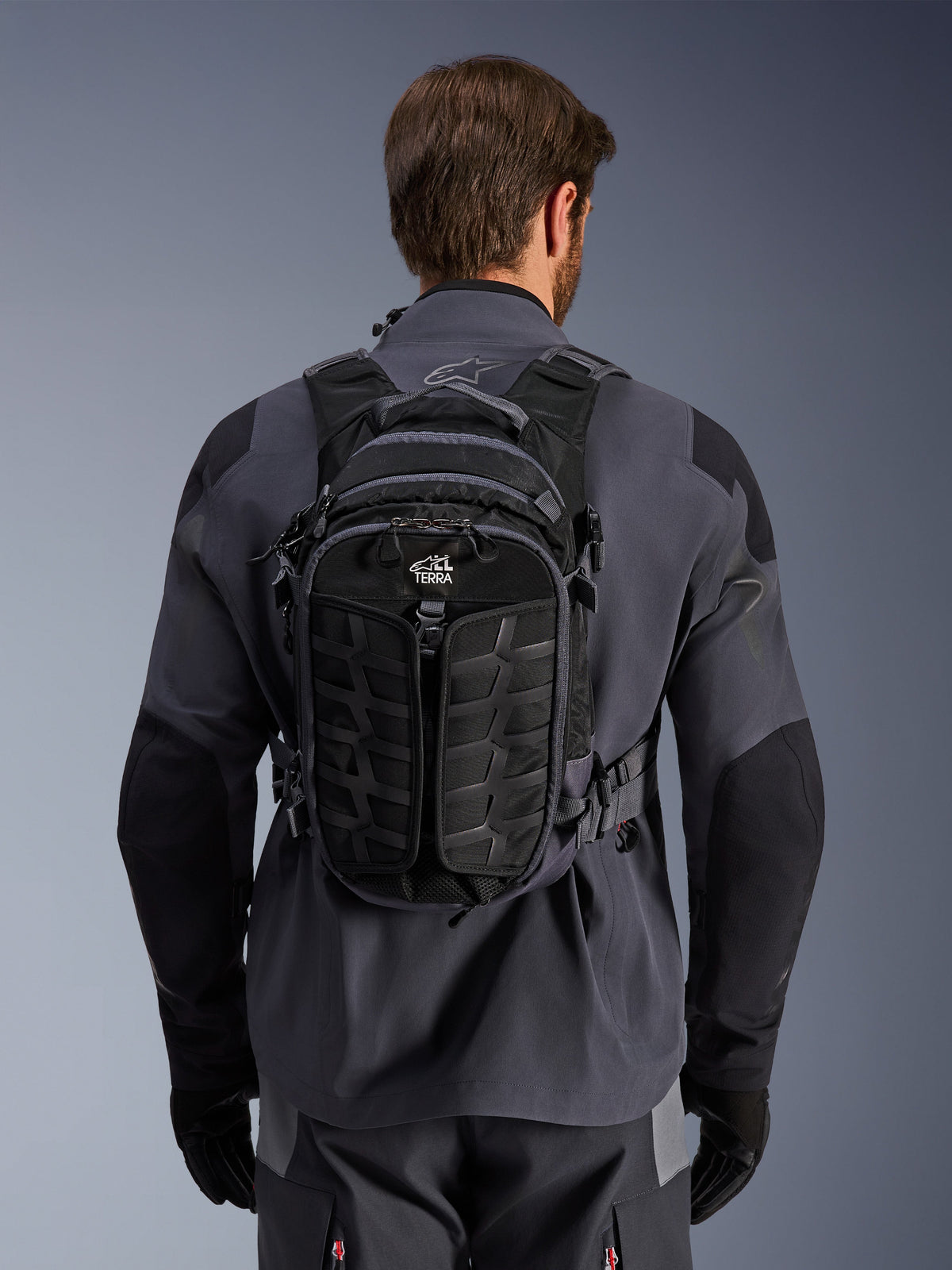 Mochila Alpinestars AT-15, mochila de motociclismo de aventura, preto e cinzento escuro, usada por uma pessoa sobre um casaco técnico de condução cinzento, vista traseira com a marca 'ALL TERRA', design ergonómico com painéis texturizados reforçados e correias seguras.