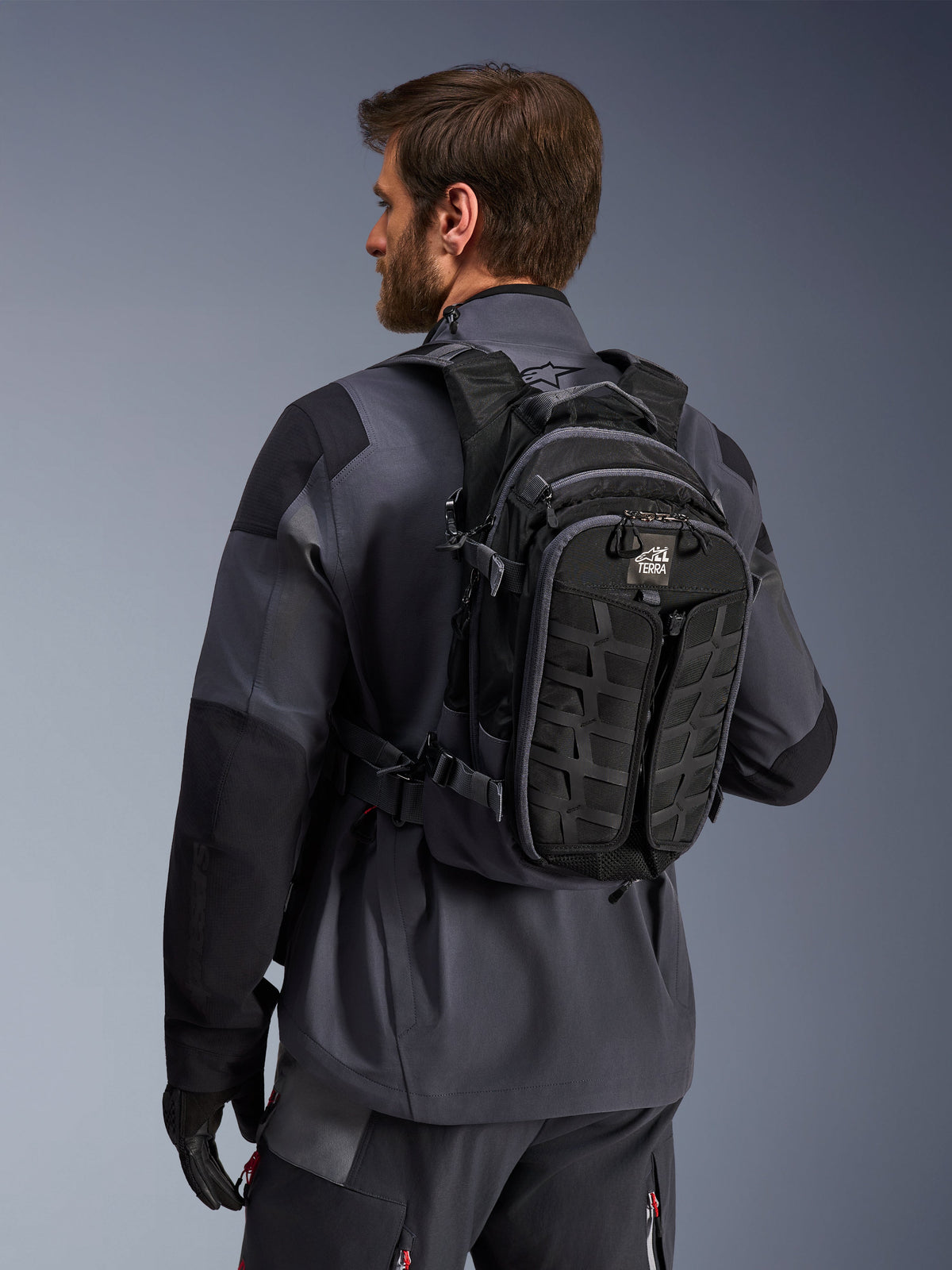 Mochila Alpinestars AT-15, mochila de motociclismo de aventura, preta e cinzento escuro, usada por um condutor com um casaco cinzento, apresentando um design técnico com sistema MOLLE e marca 'TERRA', equipamento ergonómico para todo-o-terreno
