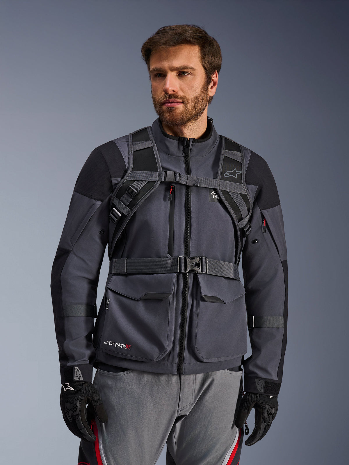 Sistema de arnês da mochila Alpinestars AT-15, Black Deep Gray, vista frontal usada por um motociclista sobre um casaco de motociclismo cinzento, com fecho magnético no peito e correia de cintura segura, combinada com luvas pretas para motociclismo de aventura
