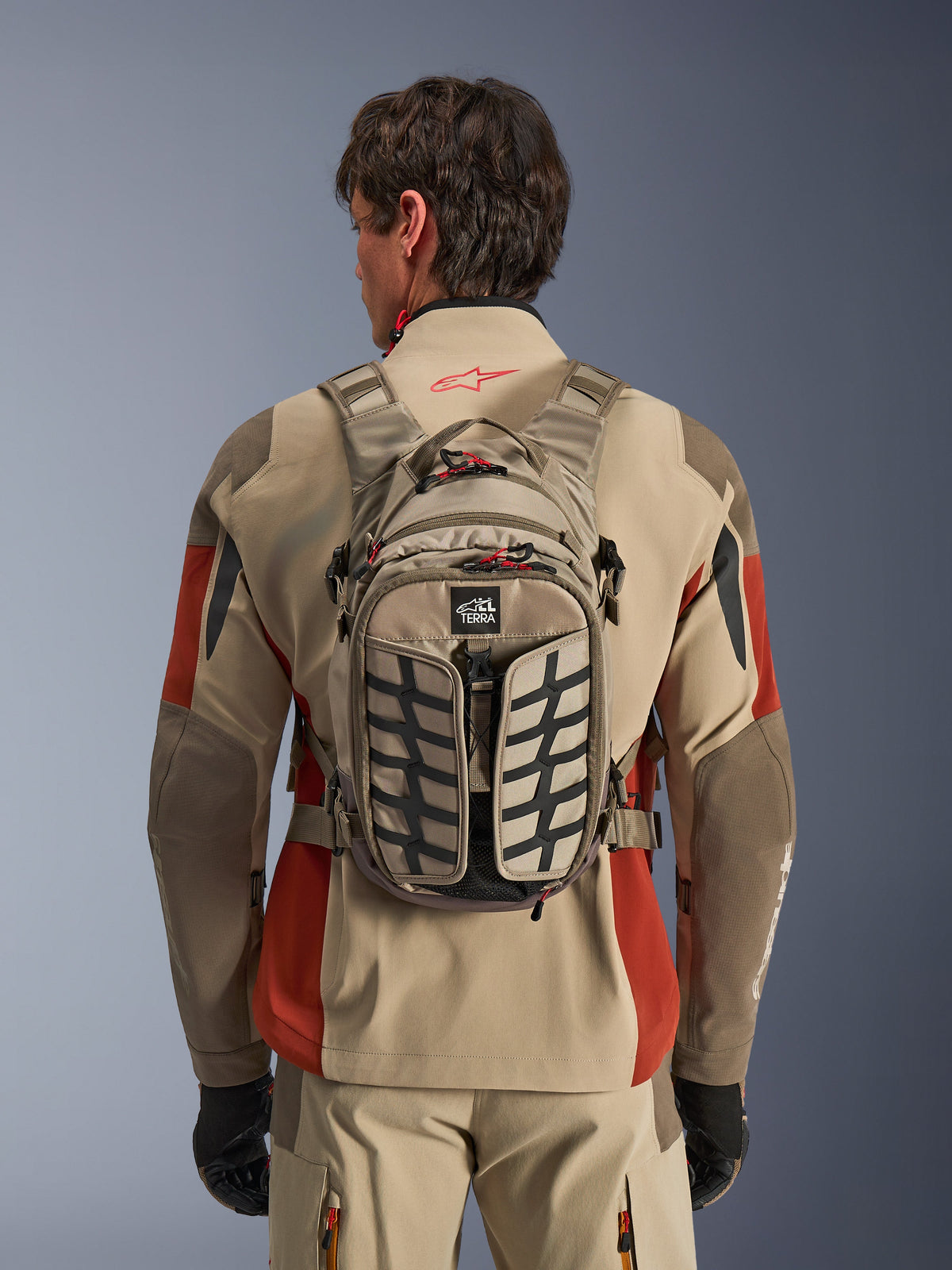 Alpinestars AT-15 Back Pack, Aluminum Walnut, mochila técnica de motociclismo de aventura usada por um piloto com casaco bege e vermelho, com padrão de rasto de pneu, logótipo TERRA, puxadores de fecho vermelhos e alças ajustáveis
