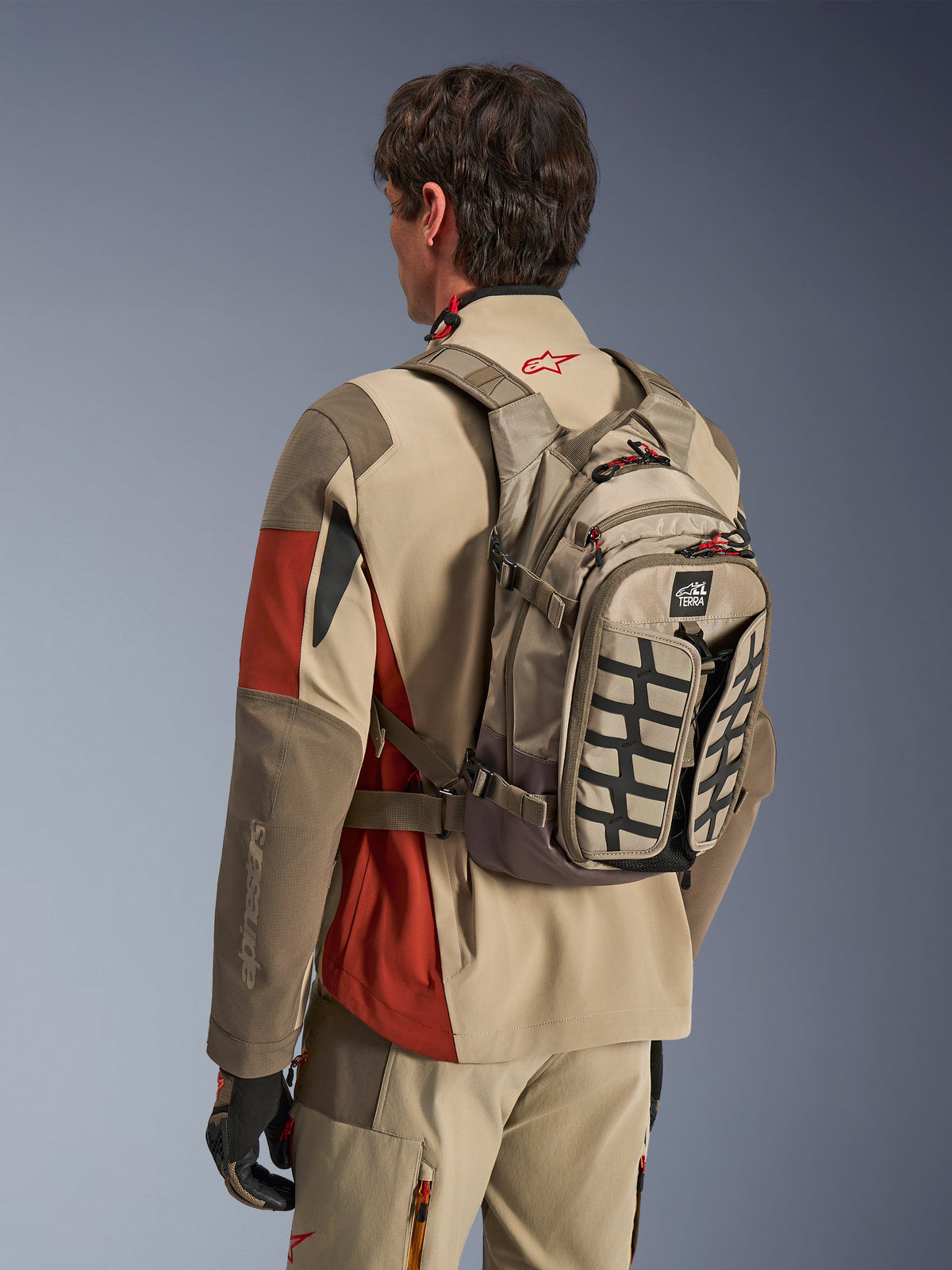 Alpinestars AT-15 Back Pack em Aluminum Walnut, mochila técnica de motociclismo usada por um piloto com equipamento de aventura a condizer, com construção têxtil bege e castanha, detalhes estruturais pretos e puxadores de fecho vermelhos, concebida para condução de aventura e todo-o-terreno.