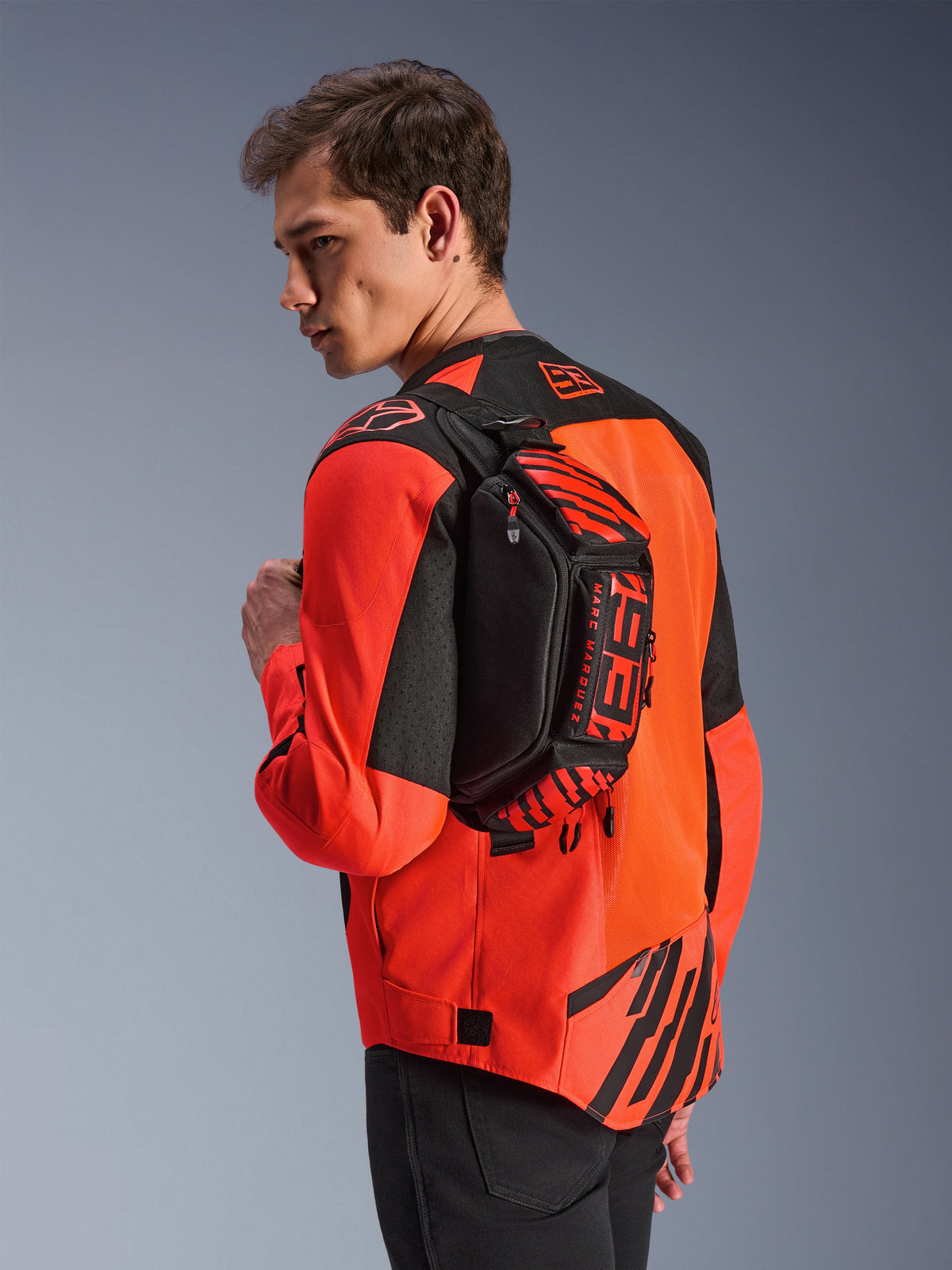 Alpinestars MM93 Waist Bag, bolsa de cintura preta com detalhes em vermelho fluo e vermelho vivo, usada por um modelo atravessada nas costas sobre um casaco de motociclismo vermelho e preto, com logótipos MM93 em destaque e grafismos de Marc Márquez