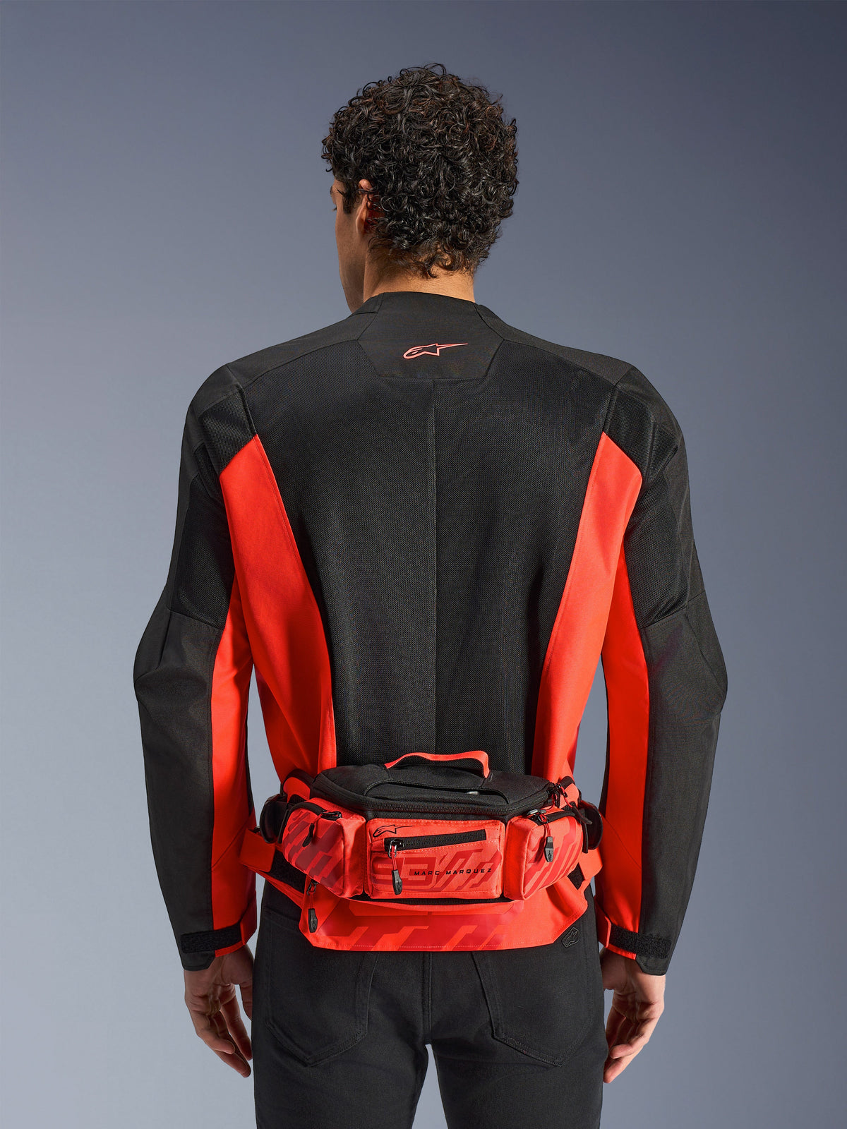 Alpinestars MM93 Waist Bag, bolsa de cintura vermelha e preta, usada sobre um casaco de motociclismo preto e vermelho, com branding Marc Marquez 93, cinto ajustável e compartimentos com fecho de correr, acessório funcional inspirado nas corridas