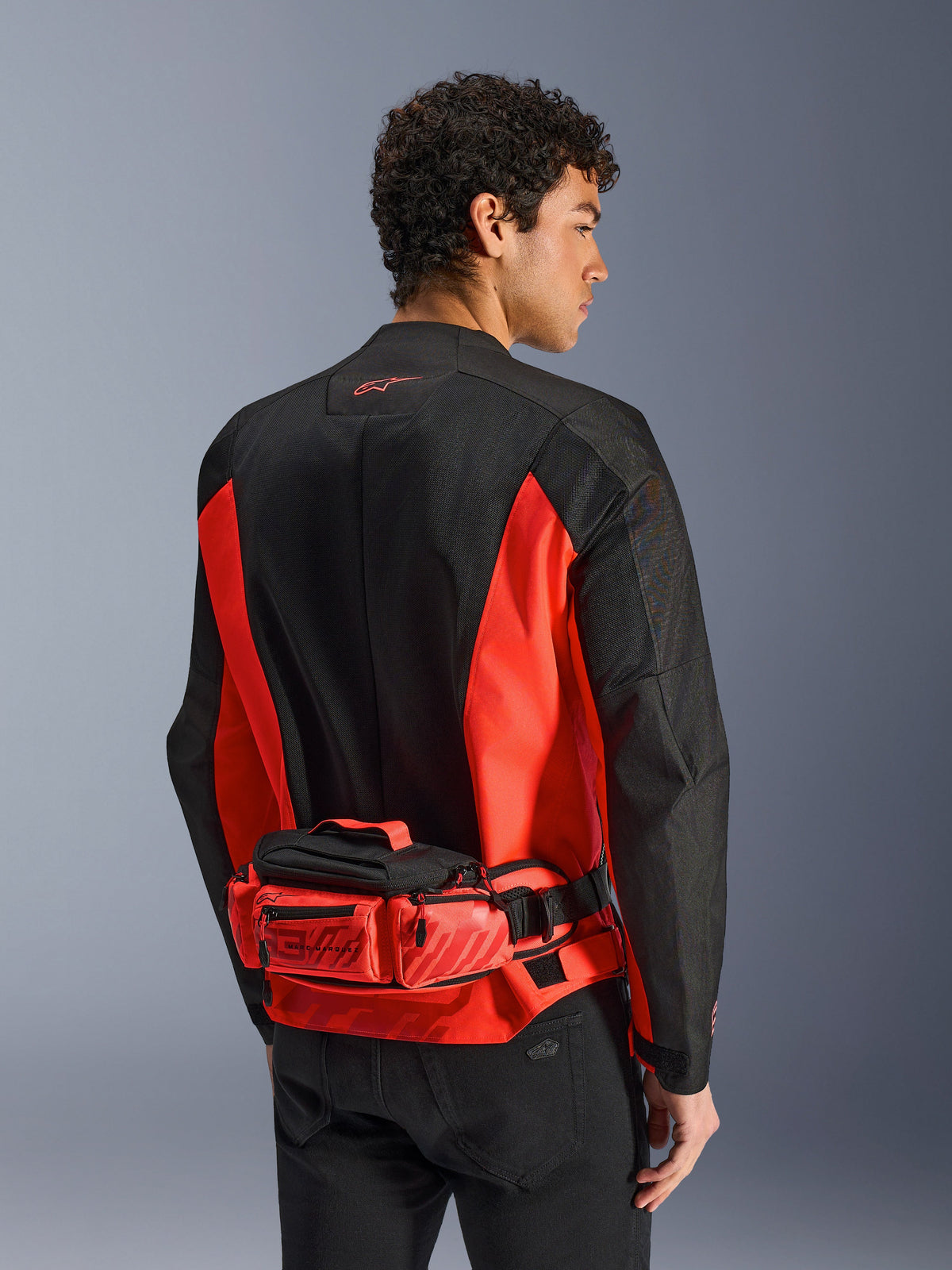 Alpinestars MM93 Waist Bag, bolsa de cintura para motociclismo, vermelho vivo e preto, usada sobre um casaco de motociclismo a condizer em preto e vermelho, com compartimentos com fecho de correr e logótipo MM93 de Marc Márquez, construção em têxtil resistente para motociclismo
