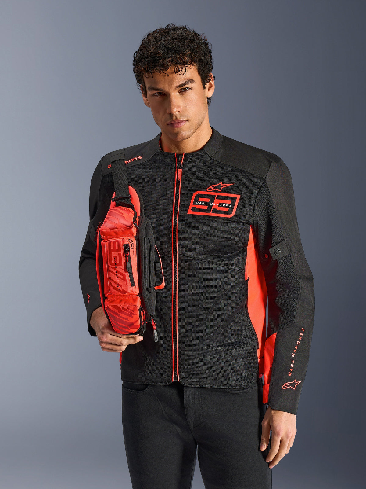Homem a usar a Alpinestars MM93 Waist Bag, em vermelho fluo e preto, usada como mala tiracolo sobre um blusão de motociclismo preto e vermelho a condizer, com o logótipo Marc Marquez 93 e design desportivo de competição