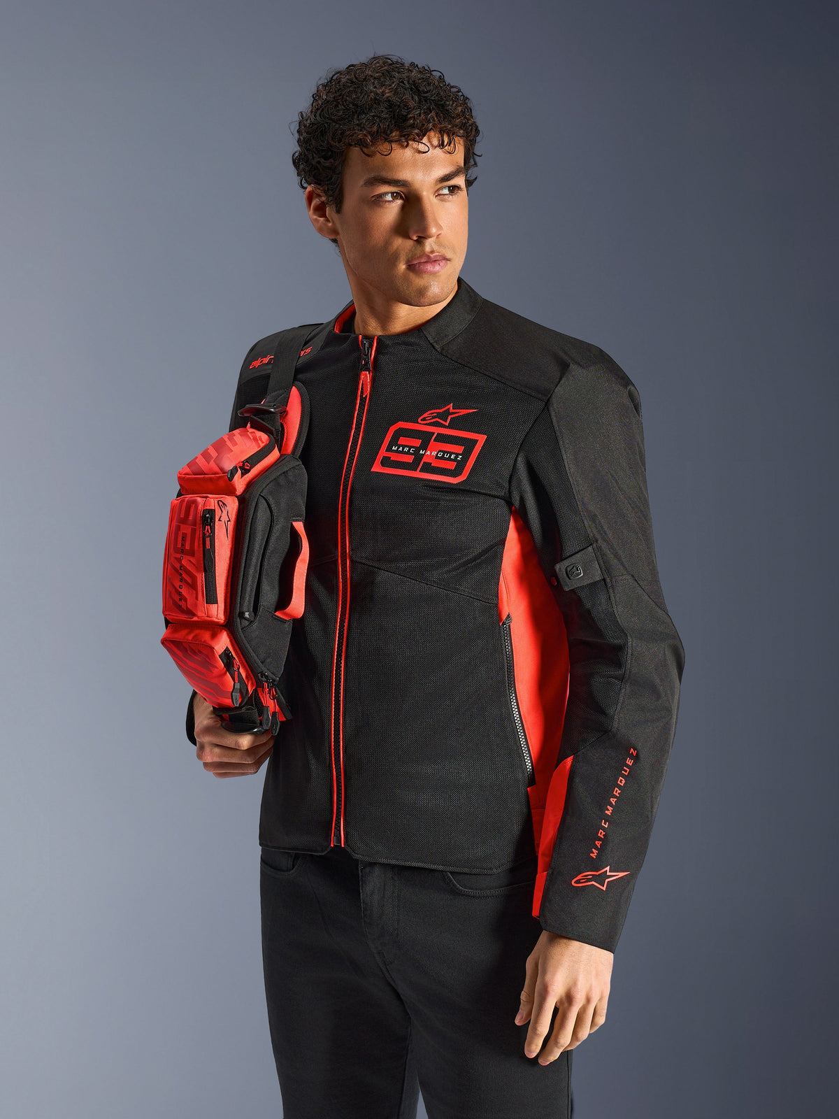 Alpinestars MM93 Waist Bag, bolsa de cintura em Vermelho Fluo e Preto, segurada por um modelo a usar um casaco de motociclismo preto e vermelho a condizer, com vários compartimentos com fecho e marca Marc Marquez, equipamento prático para condução desportiva