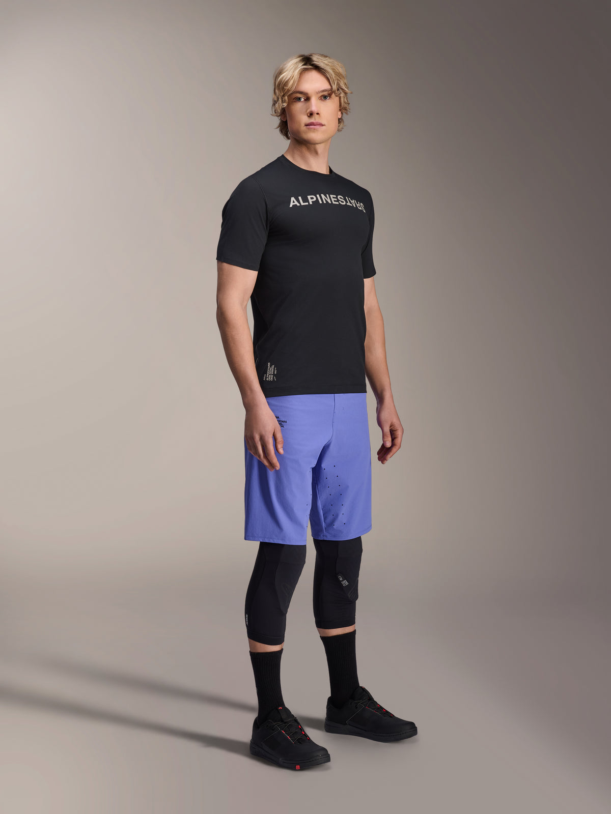 Modelo masculino a usar equipamento de mountain bike Alpinestars, com a camisola preta A-Aria Switch SS e os calções A-Aria Lite em Baja Blue, combinados com joelheiras pretas, meias pretas e sapatos de mountain bike pretos, numa fotografia de estúdio de corpo inteiro.