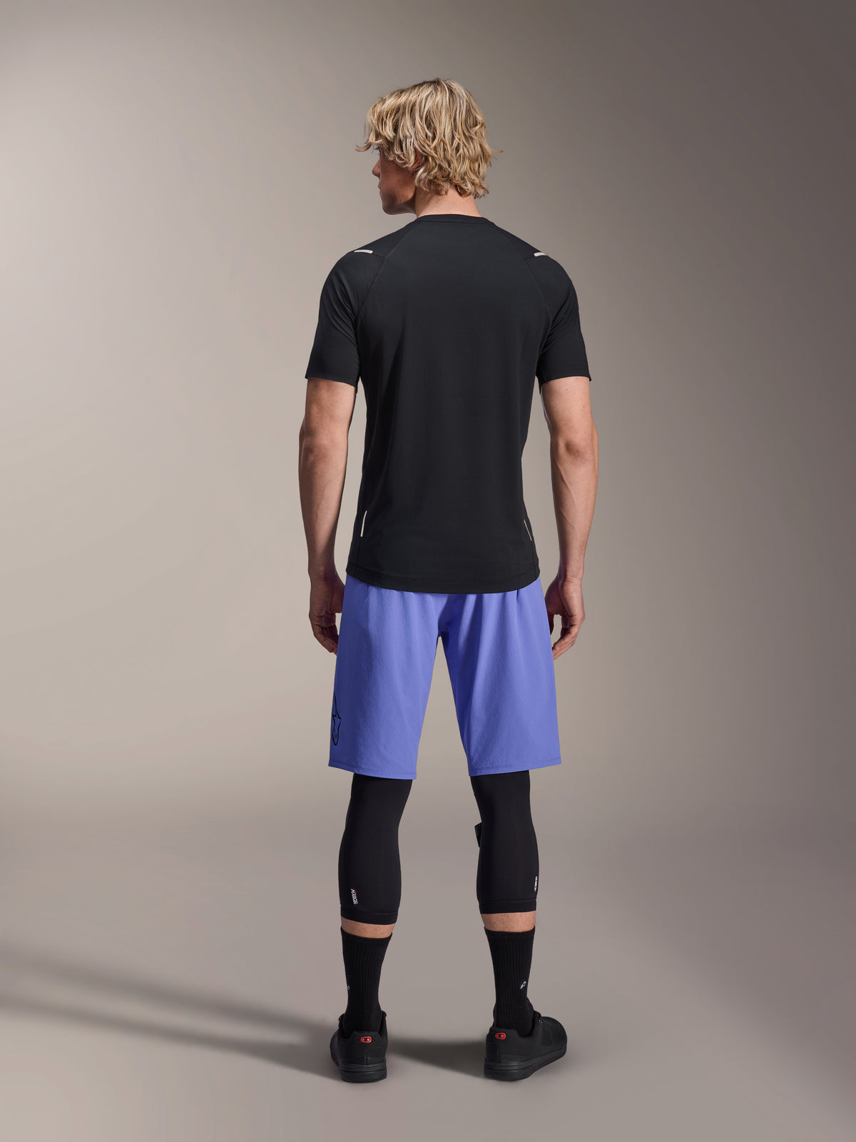 Ciclista masculino a usar calções Alpinestars A-Aria Lite, calções de MTB, Baja Blue, vista traseira, combinados com camisola de manga curta preta, joelheiras pretas e sapatos de ciclismo pretos, vestuário de performance para mountain bike
