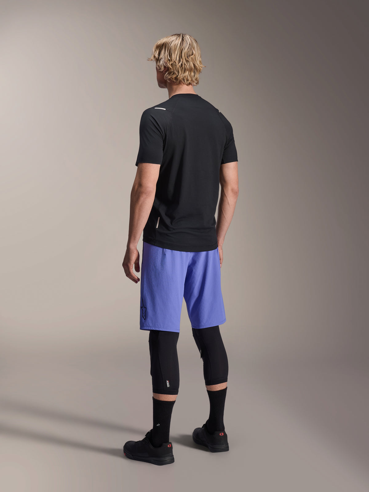Modelo masculino visto de costas a usar calções Alpinestars A-Aria Lite em Baja Blue, combinados com uma camisola preta de manga curta e joelheiras pretas, vestuário de ciclismo MTB profissional para trilhos.