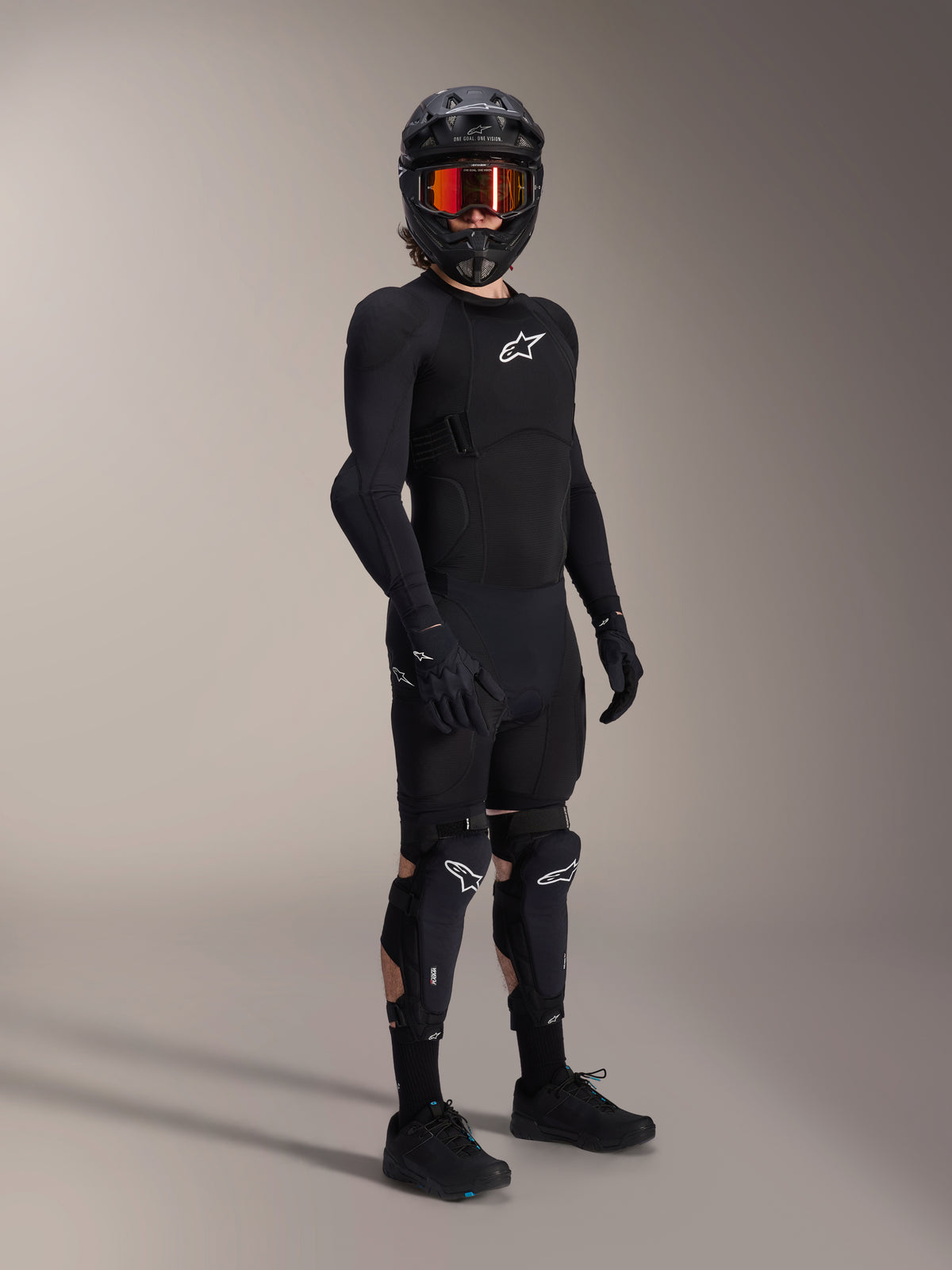 Protetores de Joelho e Canela Alpinestars A-Impact Plasma Pro, pretos e brancos, usados por um ciclista com equipamento de proteção completo para mountain bike, incluindo camisola blindada preta de manga comprida, luvas pretas e capacete integral com óculos cor de laranja