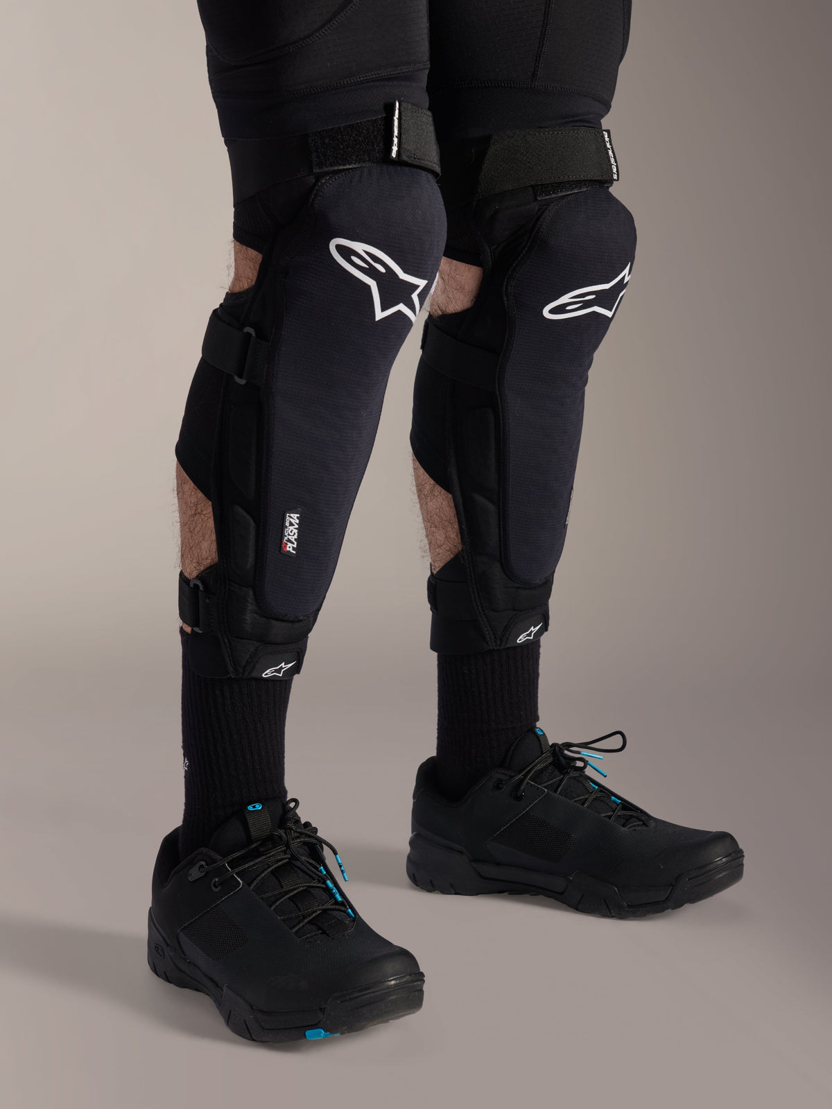 Joelheiras Alpinestars A-Impact Plasma Pro, pretas e brancas, com a marca da tecnologia Nucleon PLASMA, usadas com meias pretas e sapatos de BTT pretos, tiras de ajuste seguras, equipamento de proteção ergonómico para ciclismo todo-o-terreno