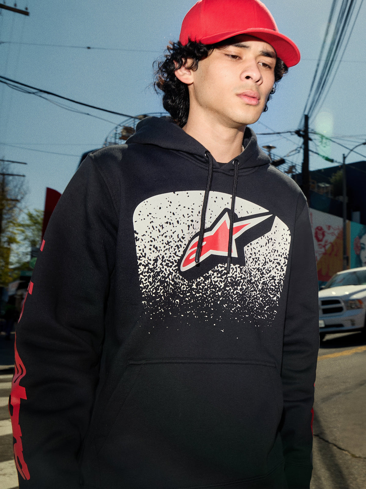 Alpinestars Obsessed Hoodie, camisola com capuz preta em fleece, gráfico de peito branco com gradação MX e logótipo de estrela vermelho, logótipos vermelhos nas mangas, streetwear casual inspirado no desporto motorizado usado num ambiente urbano