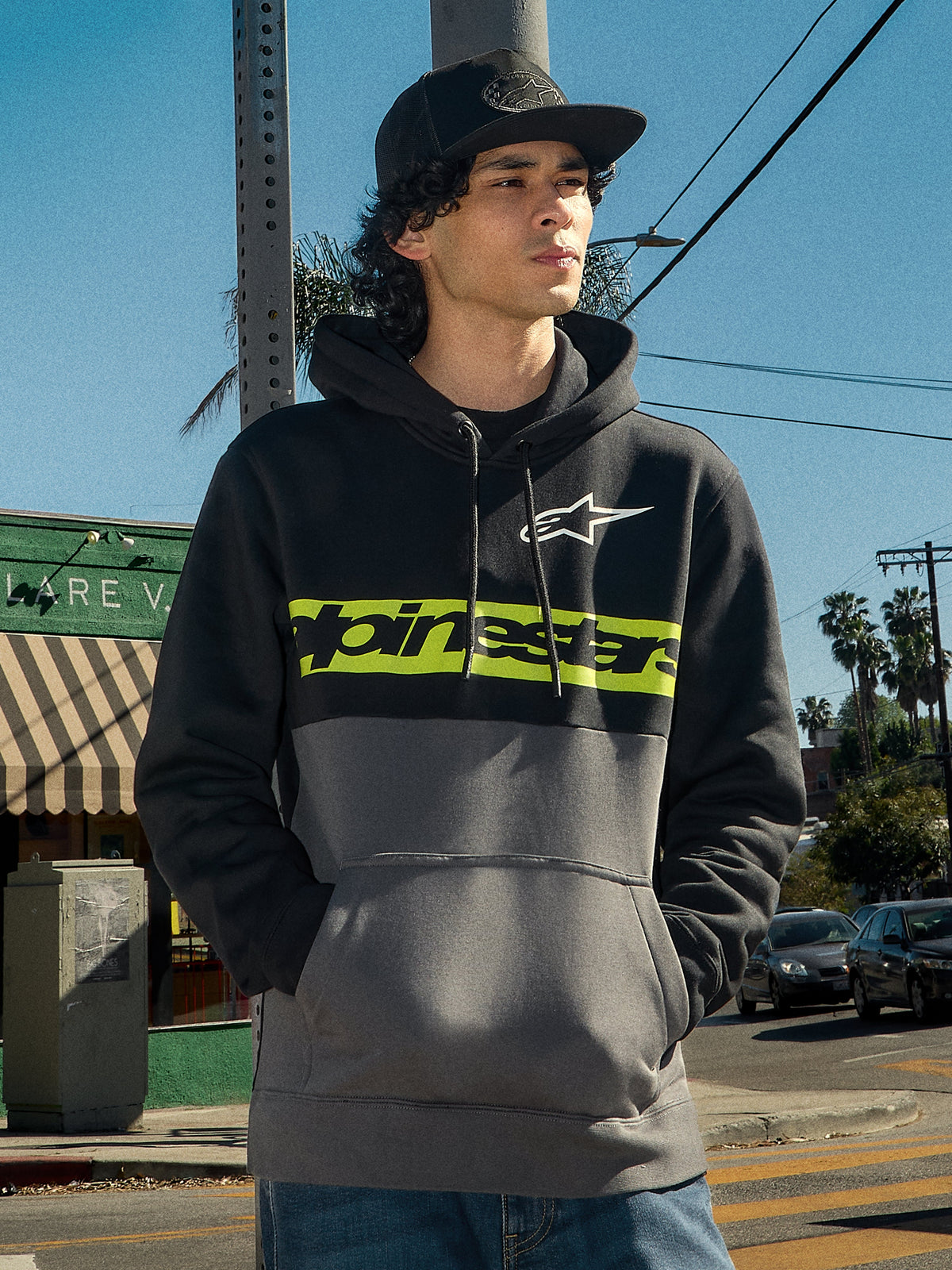 Alpinestars Parallel Hoodie, camisola com capuz streetwear lifestyle, design em blocos de cor preto e gunmetal, faixa no peito em verde lima com logótipo Alpinestars, logótipo Astars branco, capuz com cordões, usada por um homem num cenário urbano ao ar livre