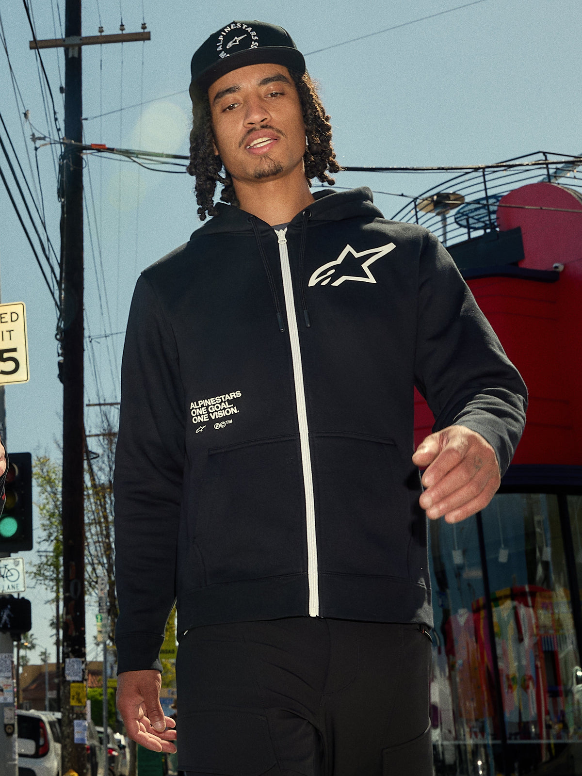 Alpinestars Transverse Hoodie, camisola de fleece preta com fecho frontal, fecho de correr branco e logótipo Astars branco no peito, impressão de texto 'One Goal One Vision', estilo casual streetwear, usada por um homem num ambiente urbano exterior