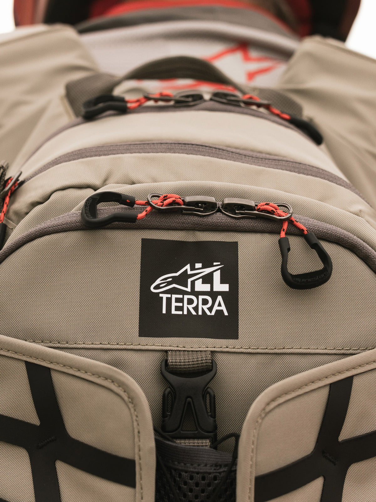 Mochila Alpinestars AT-15, Aluminum Walnut, vista de pormenor do emblema do logótipo All Terra em tecido técnico bege, com puxadores de fecho em cordão laranja e detalhes de construção robusta