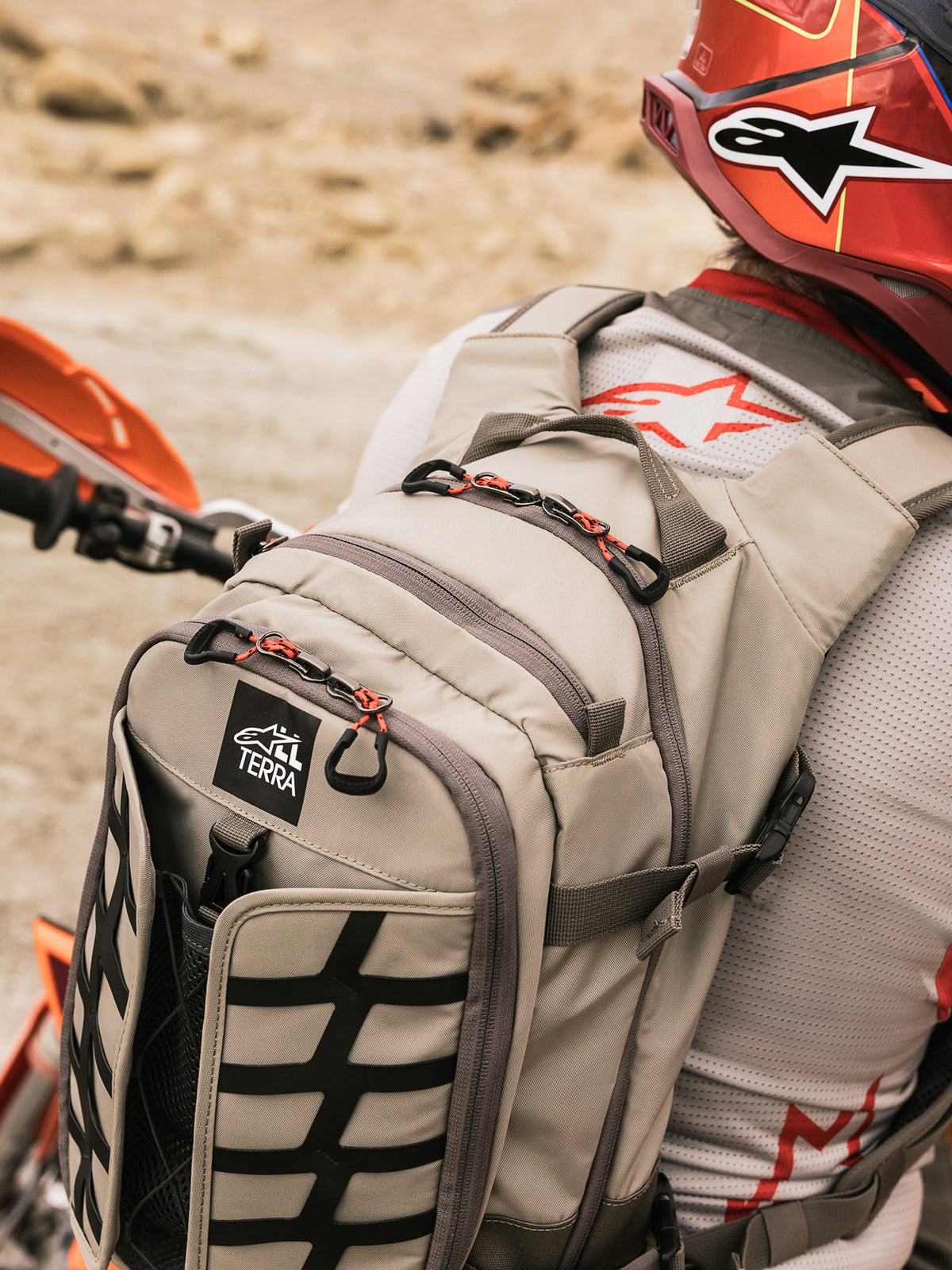 Mochila Alpinestars AT-15, mochila de motociclismo de aventura, em Aluminum Walnut e preto, usada por um piloto num cenário de deserto, com puxadores de fecho vermelhos, emblema com o logótipo TERRA e fitas externas pretas, combinada com um capacete vermelho e camisola Alpinestars branca.