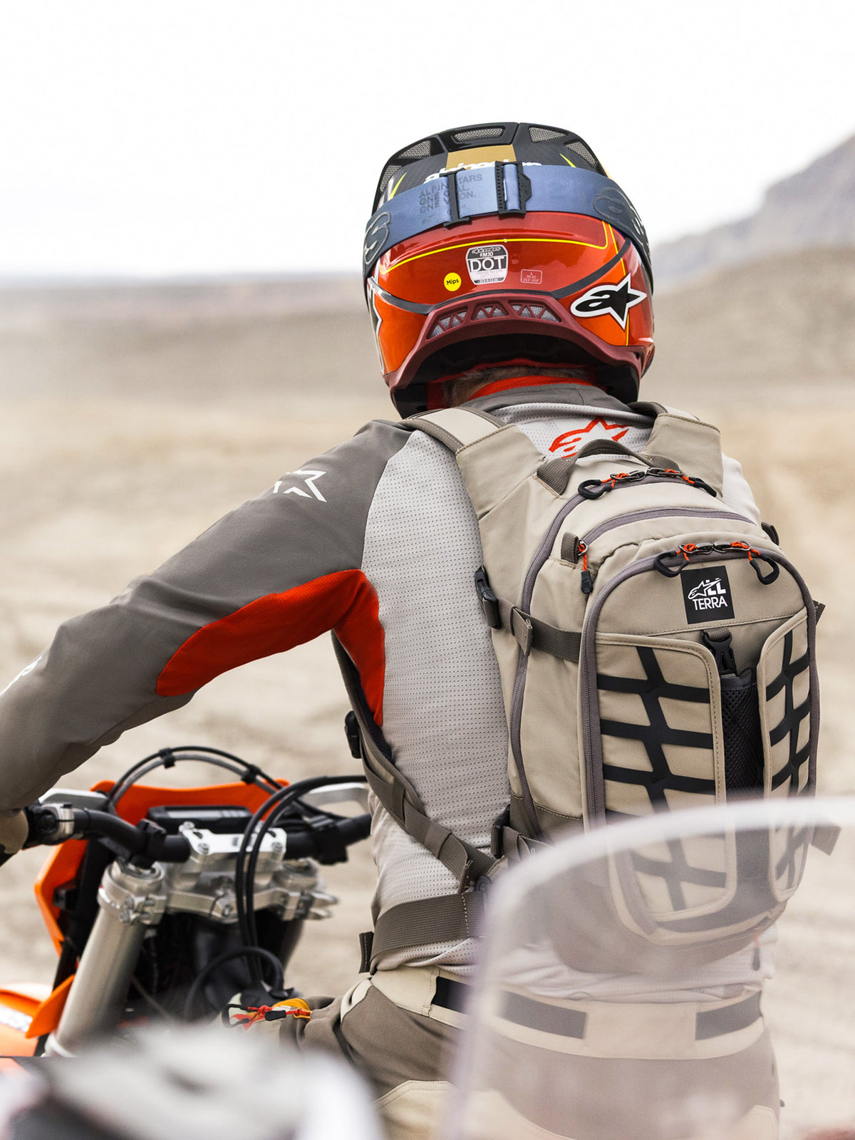 Mochila Alpinestars AT-15 em Aluminum Walnut, utilizada por um motociclista de aventura numa paisagem desértica. Apresenta uma construção têxtil bege robusta com fitas pretas, detalhes de fechos em laranja e marca 'All Terra'. Combinada com um capacete vermelho e laranja e equipamento de condução cinzento para touring fora de estrada.