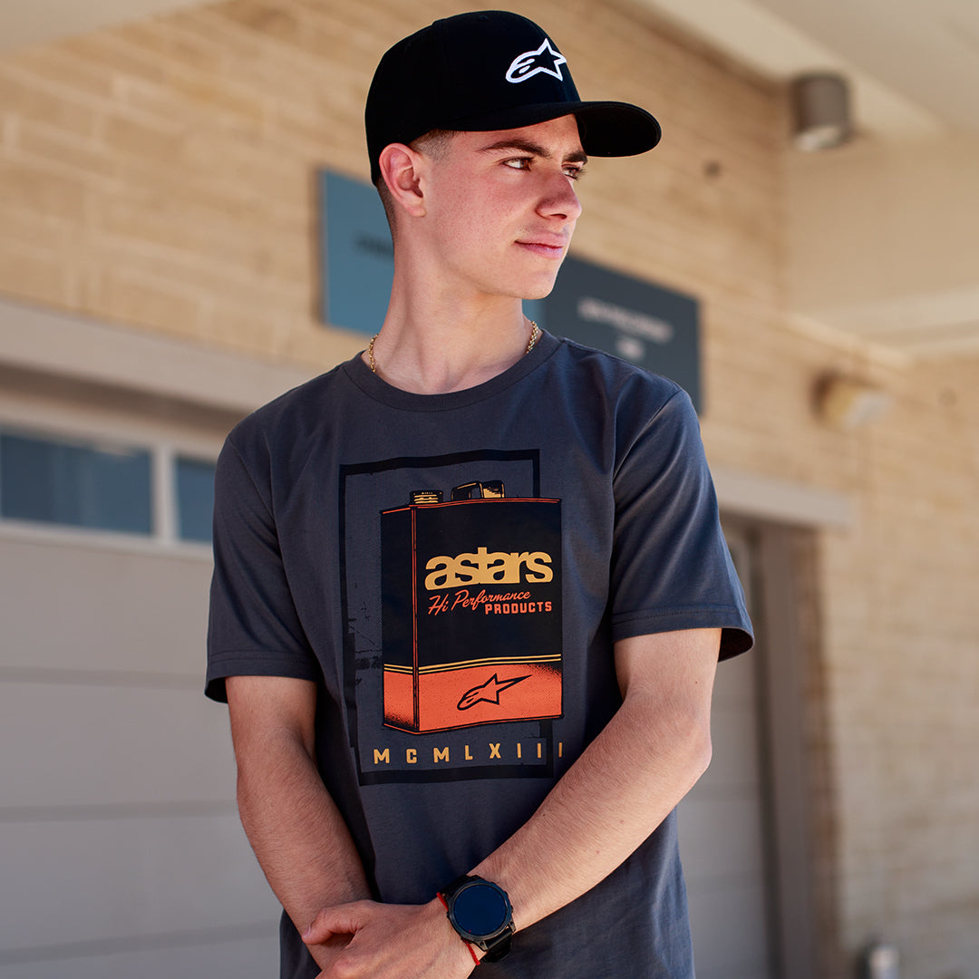 Alpinestars Galun CSF Tee, t-shirt de manga curta cinzenta, grafismo frontal de uma lata de gasolina vintage com a marca 'astars' e numeração romana 'MCMLXIII', vestida por um homem com um boné preto com logótipo, estilo de vida casual de desportos motorizados.