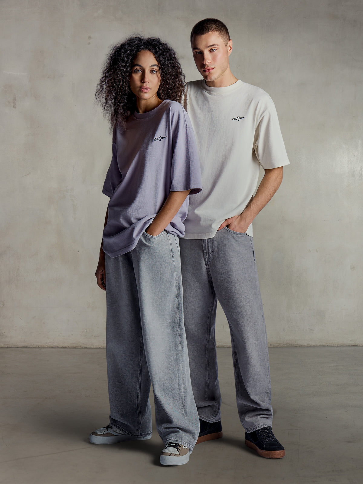 Alpinestars Essentials Ageless SS Tee, vista lifestyle de dois modelos a usar t-shirts de manga curta em roxo claro e prateado, algodão orgânico de corte relaxado com um logótipo subtil no peito, combinadas com calças de ganga largas para um estilo casual streetwear.