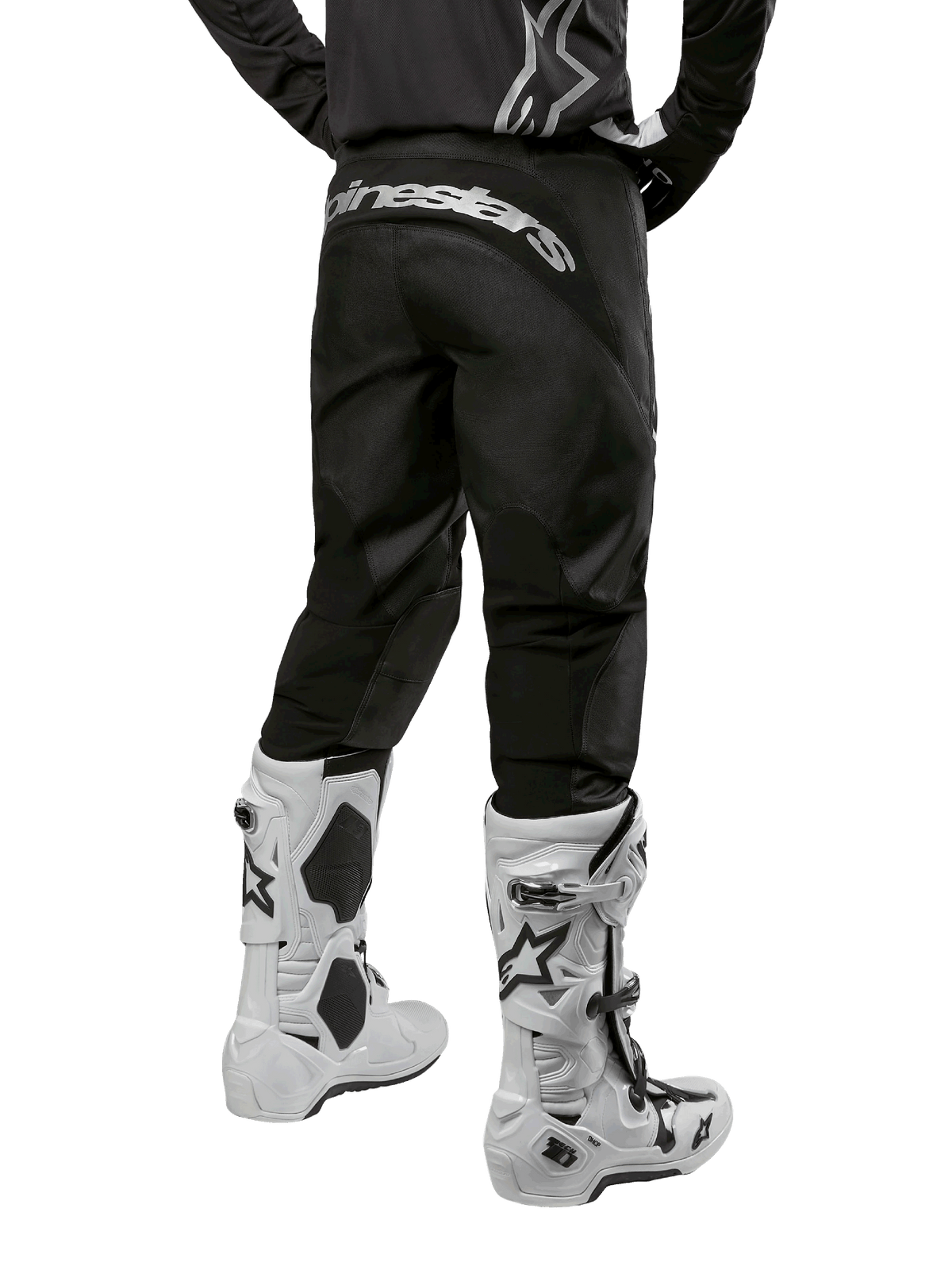 Alpinestars 2024 Fluid Graphite Pants, calças de motocross, pretas e prateadas, vista traseira com marca Alpinestars prateada na pala traseira, usadas com botas de motocross brancas e pretas, design têxtil durável para condução todo-o-terreno