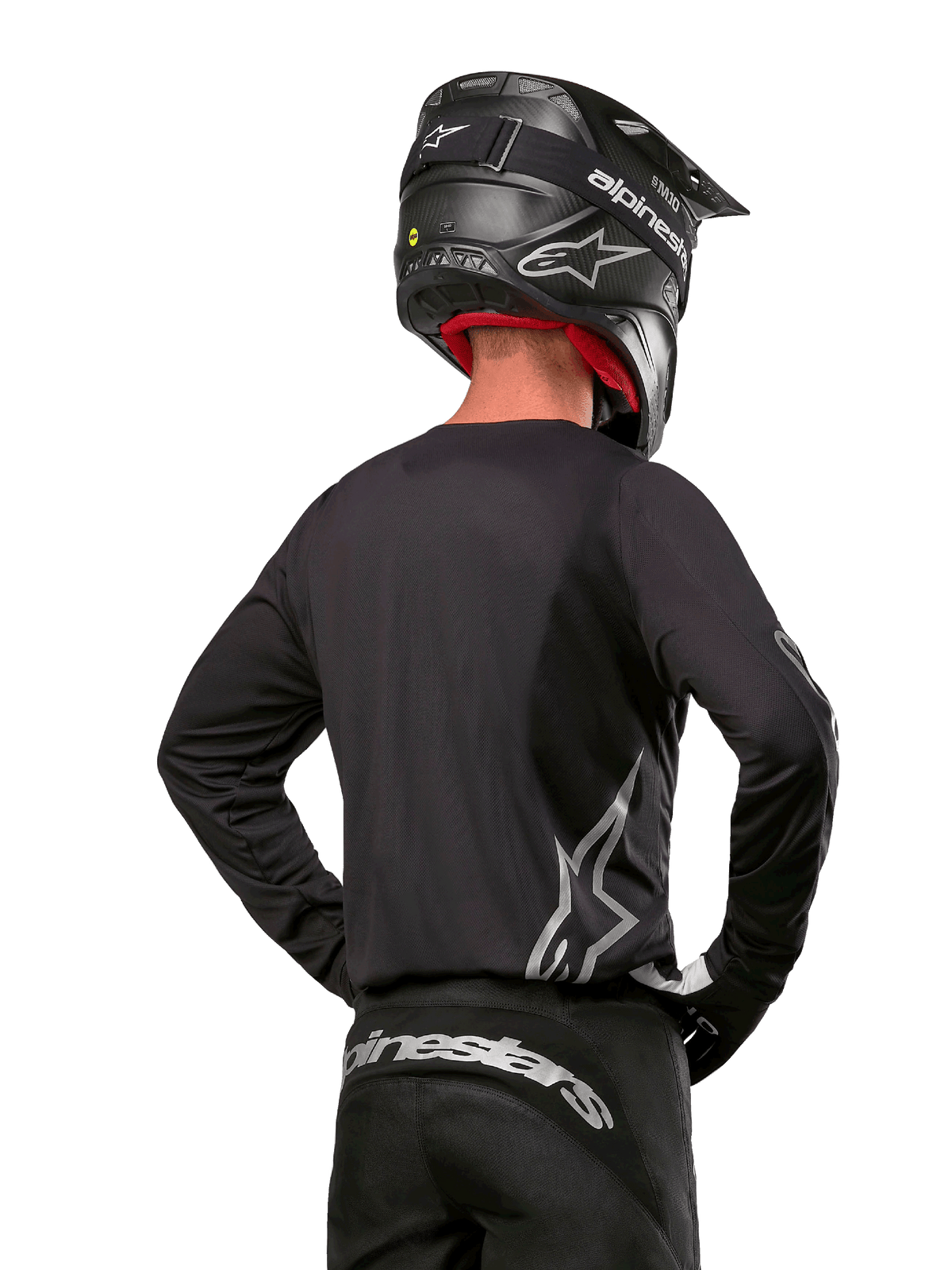 Piloto de motocross visto de costas a usar a camisola Alpinestars 2024 Fluid Graphite em preto e prateado, com logótipo prateado grande na lateral, capacete preto com textura de carbono e calças pretas, equipamento profissional de motocross