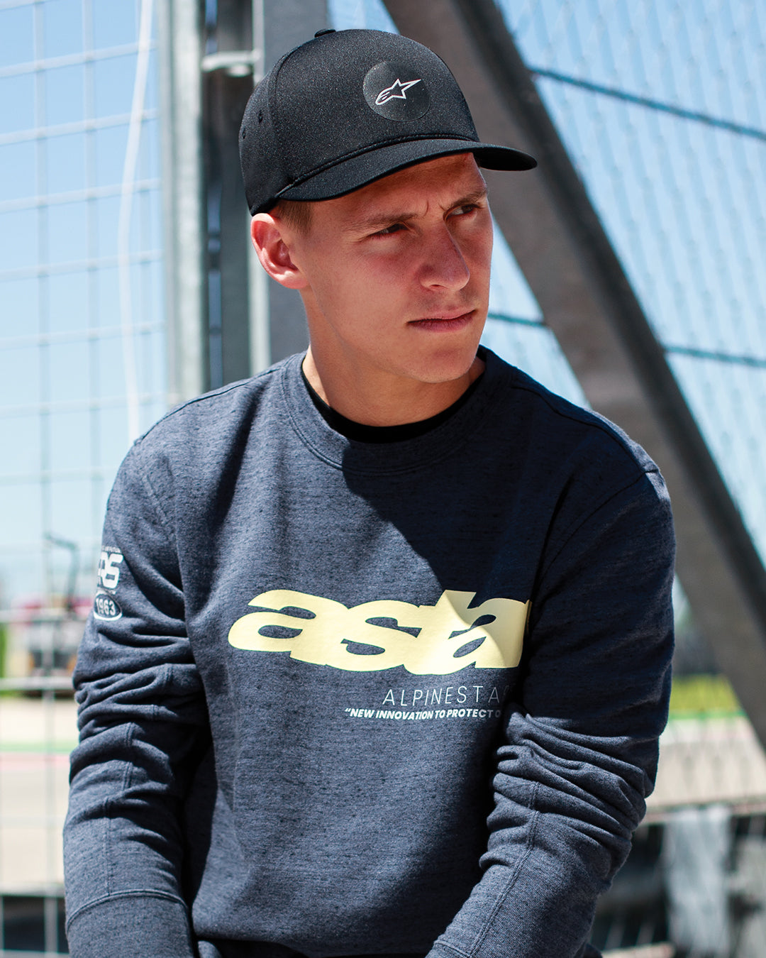 Fabio Quartararo a usar o boné Alpinestars Apex Hat em preto, boné de copa estruturada com pala curvada e logótipo circular com estrela, combinado com uma sweatshirt cinzenta escura mesclada com logótipo amarelo, estilo de vida casual inspirado no desporto motorizado.