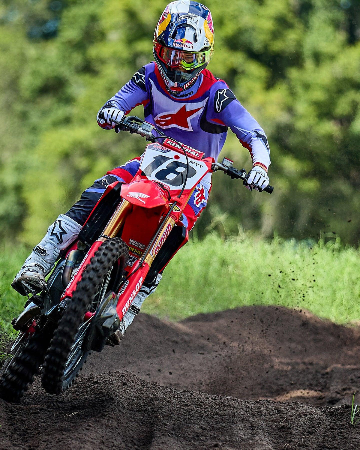 Piloto de motocross numa mota Honda vermelha, com a camisola Alpinestars 2025 Fluid Wurx em roxo e vermelho vivo, calças a condizer, botas brancas e capacete Red Bull, a percorrer uma curva numa pista de terra
