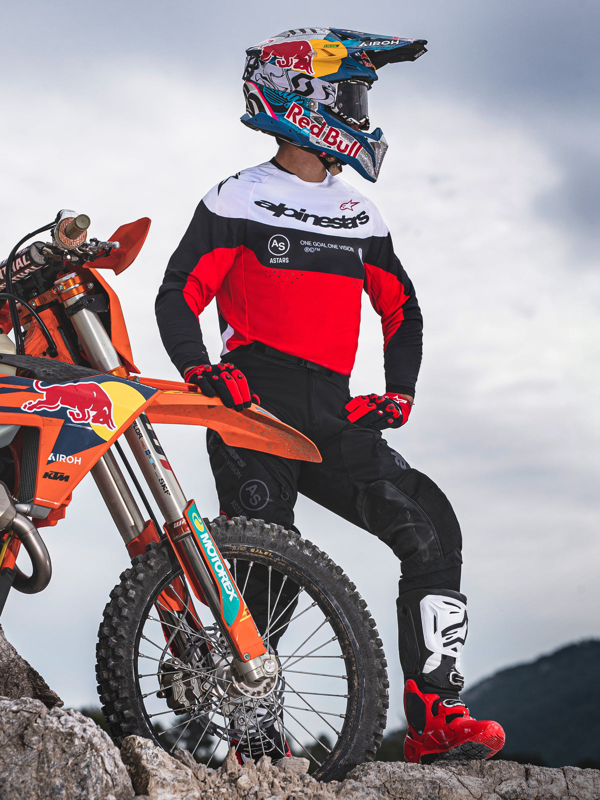 Piloto de motocross com a Camisola Alpinestars Pro-Dura em preto, vermelho vivo e branco, de pé junto a uma mota KTM em terreno rochoso, combinada com calças pretas e botas de motocross vermelhas, cenário de enduro ao ar livre.