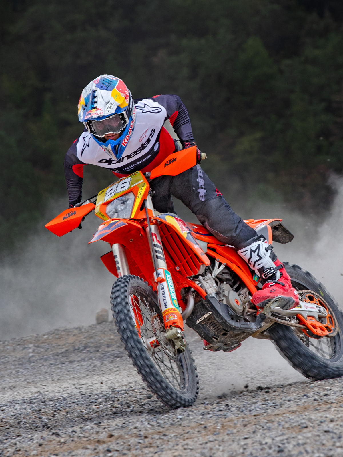 Piloto de motocross em ação a usar a camisola Alpinestars Pro-Dura em preto, vermelho vivo e branco, a conduzir uma mota de terra cor de laranja num trilho de off-road poeirento, acompanhada por calças pretas e botas vermelhas e brancas