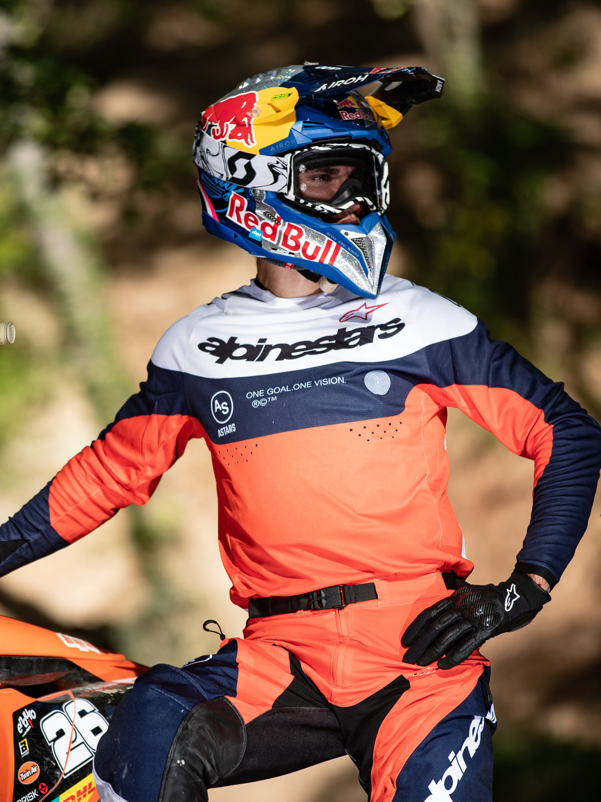 Piloto de motocross a usar a camisola Alpinestars Pro-Dura em azul-marinho, laranja vibrante e branco, com logótipo de destaque no peito e perfurações a laser, combinada com um capacete de corrida Red Bull, equipamento profissional para performance em enduro e todo-o-terreno