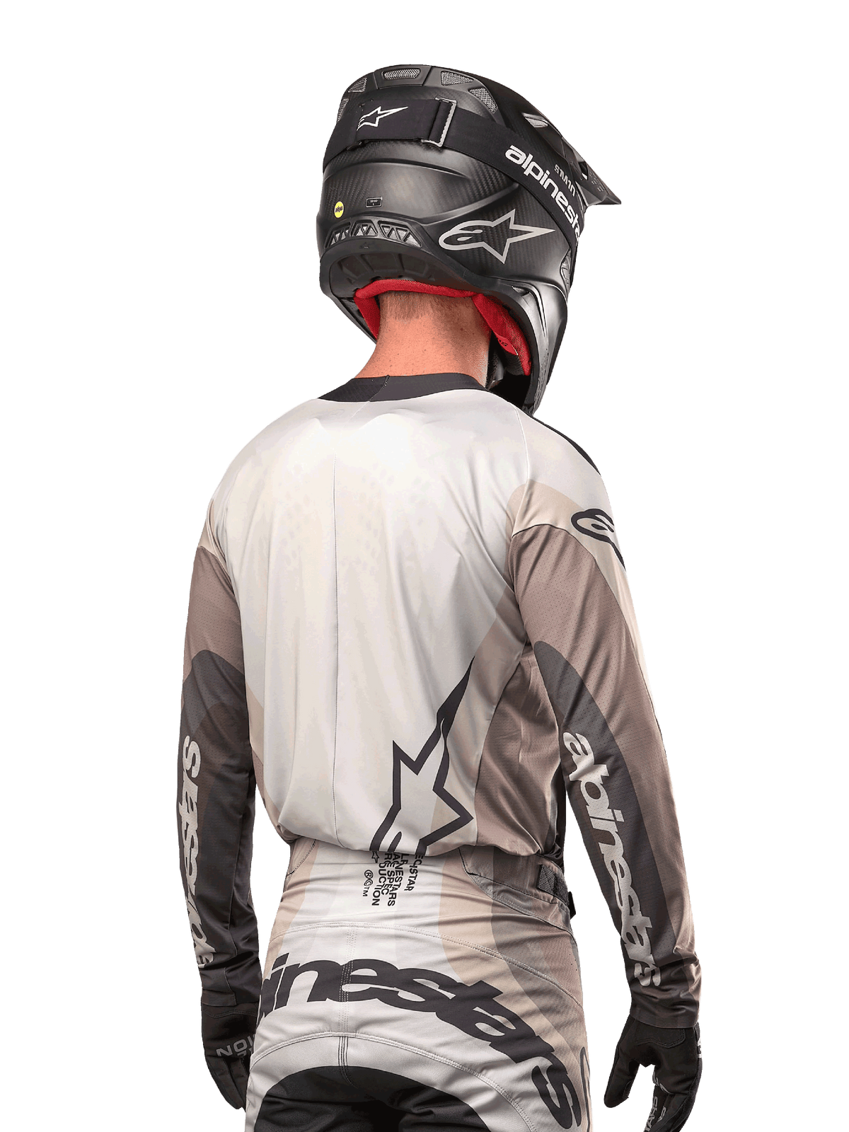 Piloto de motocross visto de trás a usar a camisola Alpinestars Techstar Pneuma 2024 em Dark Sand Iron Dust Gray, combinada com um capacete preto com acabamento em carbono, com um logótipo de estrela grande nas costas e a marca Alpinestars nas mangas.