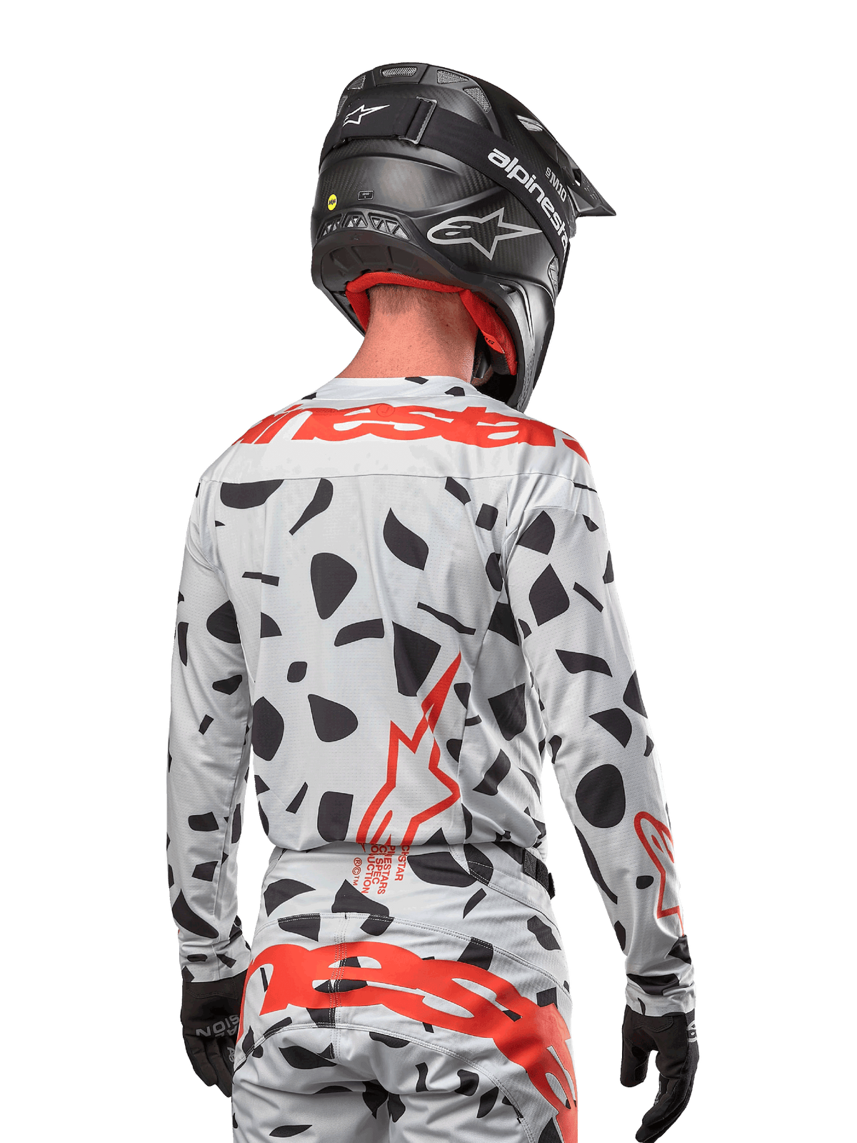 Piloto de motocross visto de costas a usar a camisola Alpinestars 2024 Techstar Rantera em Haze Gray Camo, com uma base cinza clara e manchas gráficas pretas, logótipos Alpinestars em vermelho vibrante nas costas e ombros, acompanhado por calças a condizer e capacete preto.