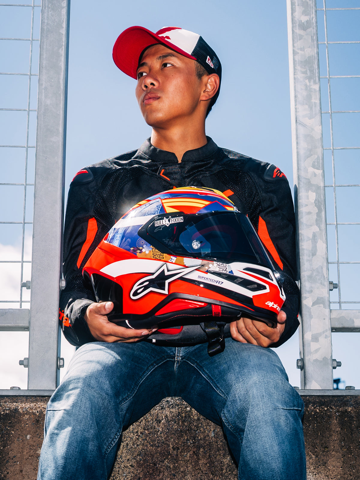 Capacete Alpinestars S-R7 Taiyo Furusato, capacete integral de corrida para moto, grafismo em vermelho, branco e azul, segurado pelo piloto Taiyo Furusato com casaco preto, vista ao ar livre junto à pista.