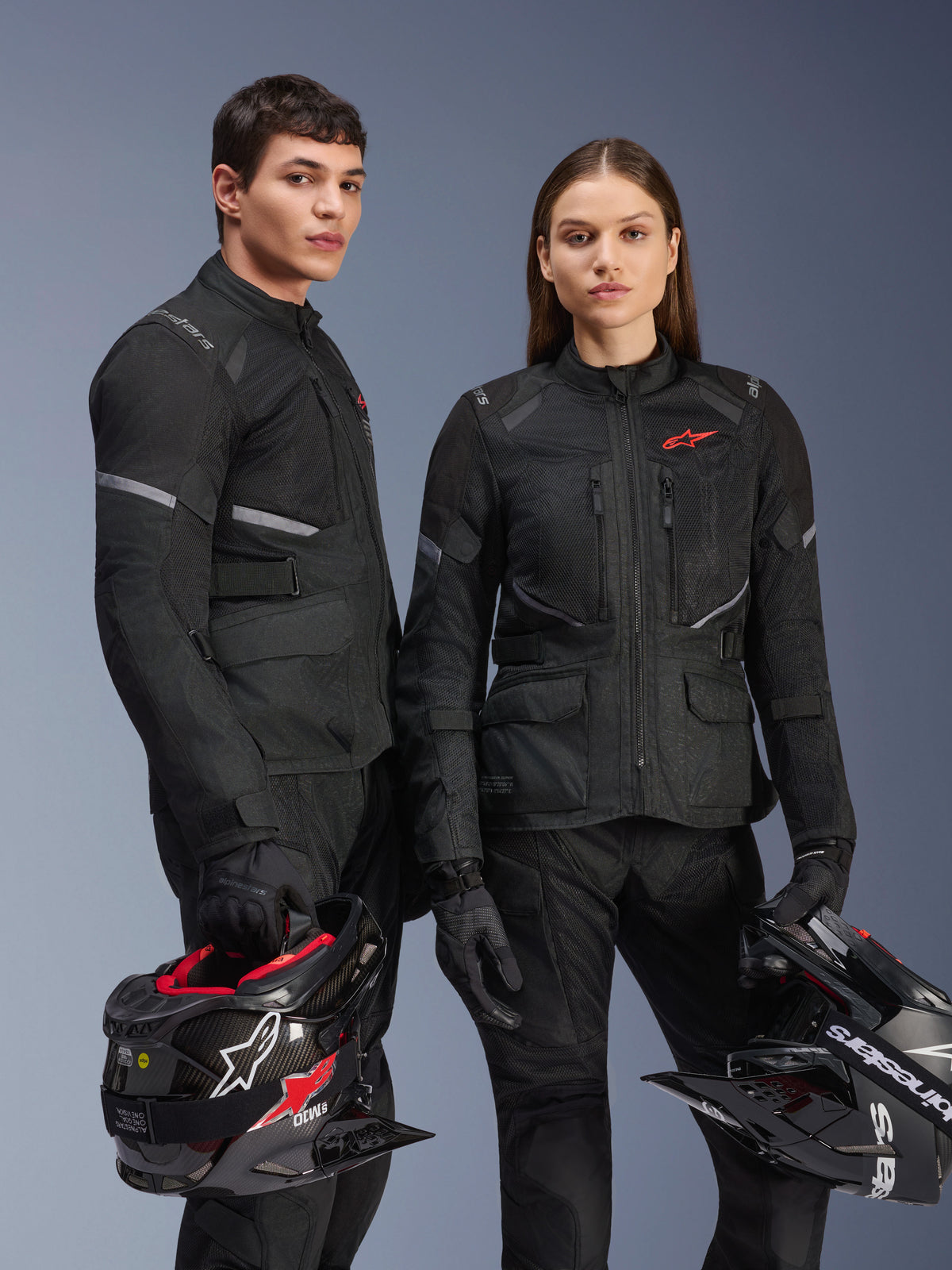 Modelos masculino e feminino a usar casacos Alpinestars Stella Andes Air Drystar® pretos, equipamento de motociclismo de aventura com logótipos de estrela vermelhos, painéis de ventilação em malha e construção têxtil técnica, segurando capacetes de aventura em estúdio