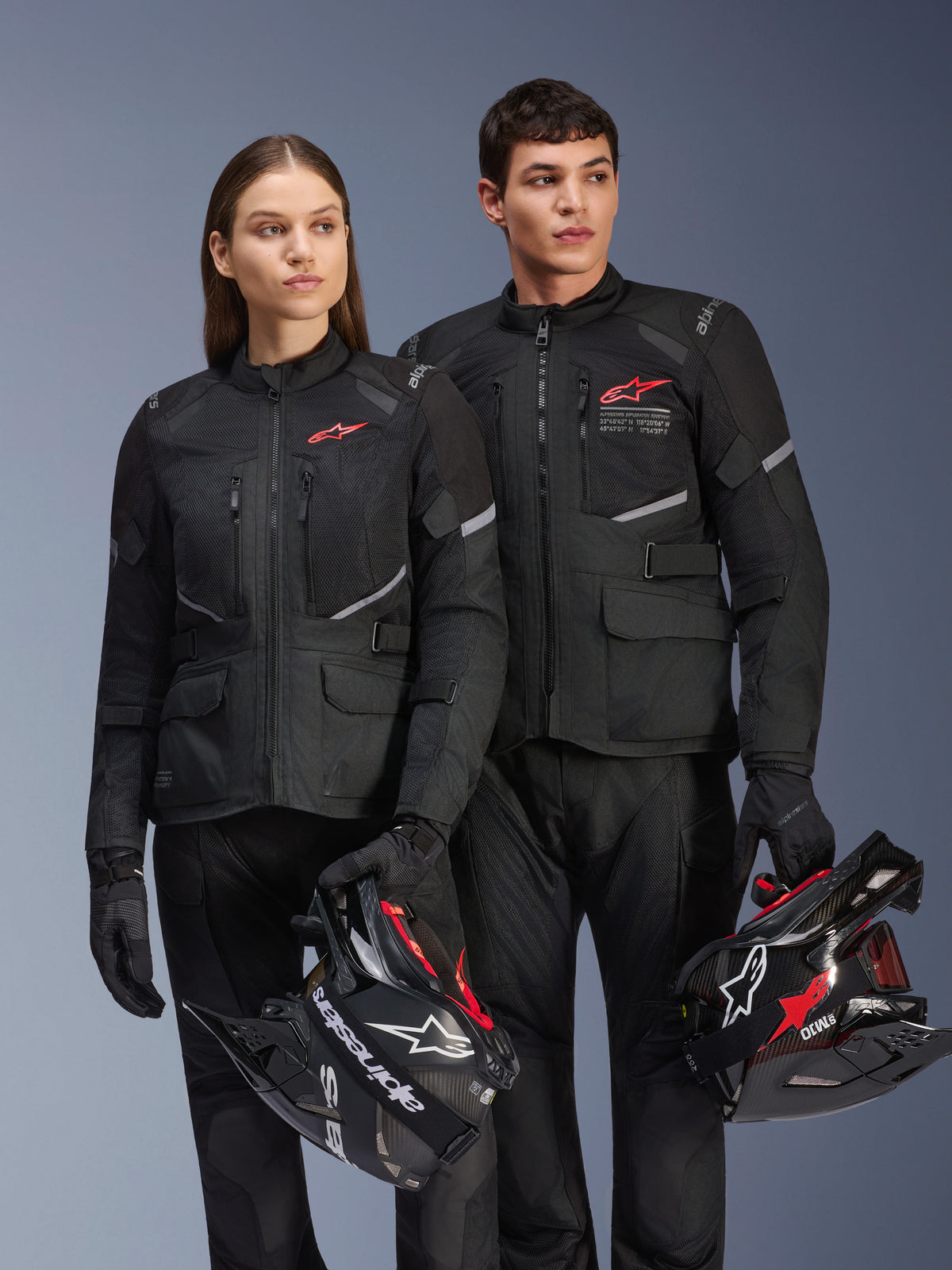 Homem e mulher a usar casacos Alpinestars Andes Air Drystar®, equipamento de touring de aventura preto com painéis de malha e detalhes refletores, calças e luvas pretas a condizer, a segurar capacetes de motocross, fotografia de estúdio de vestuário profissional de motociclismo