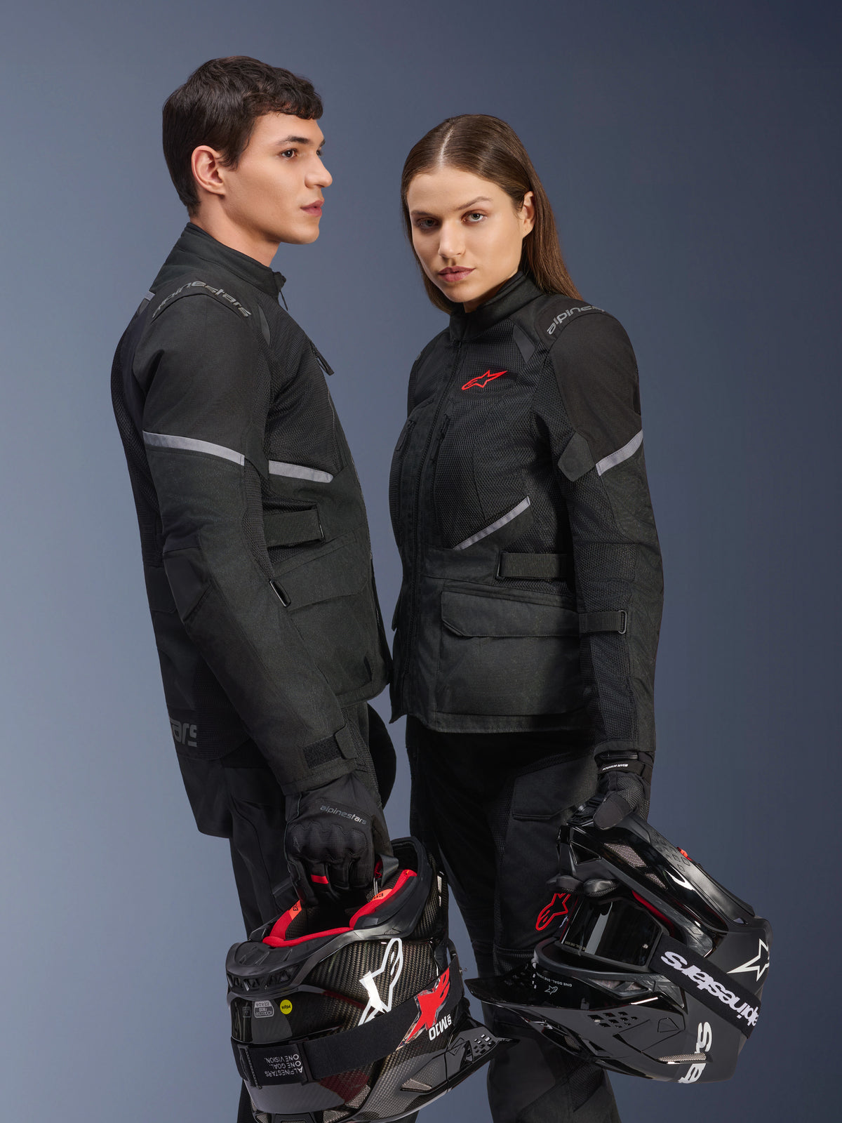 Pilotos masculino e feminino com equipamento de motociclismo Alpinestars, apresentando o Casaco Stella Andes Air Drystar® em preto, calças e luvas têxteis a condizer, ambos a segurar capacetes pretos e brancos, fotografia de estúdio de vestuário de aventura e turismo coordenado