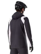Alpinestars A-Dura Rain Jacket, Mountain Bike Rain Jacket, Black with white sleeve panels, rear view of rider wearing a black MTB helmet and gloves, technical 2.5-layer waterproof construction, mountain biking apparel