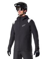 Alpinestars A-Dura Rain Jacket, Mountain Bike Rain Jacket, Black with light gray shoulder accents, worn by a rider with a black MTB helmet and sunglasses, white chest logo, waterproof construction, coordinated with black pants and gloves for off-road cycling