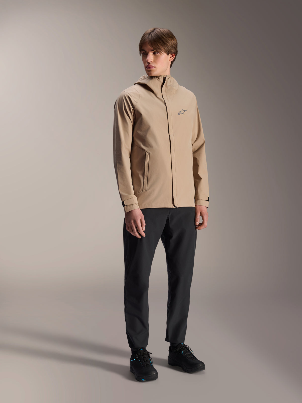 Modelo masculino a usar o Casaco de Chuva Alpinestars A-Dura em castanho claro, combinado com calças e sapatos pretos, casaco de chuva minimalista de exterior para ciclismo de montanha com design com capuz e um pequeno logótipo no peito.