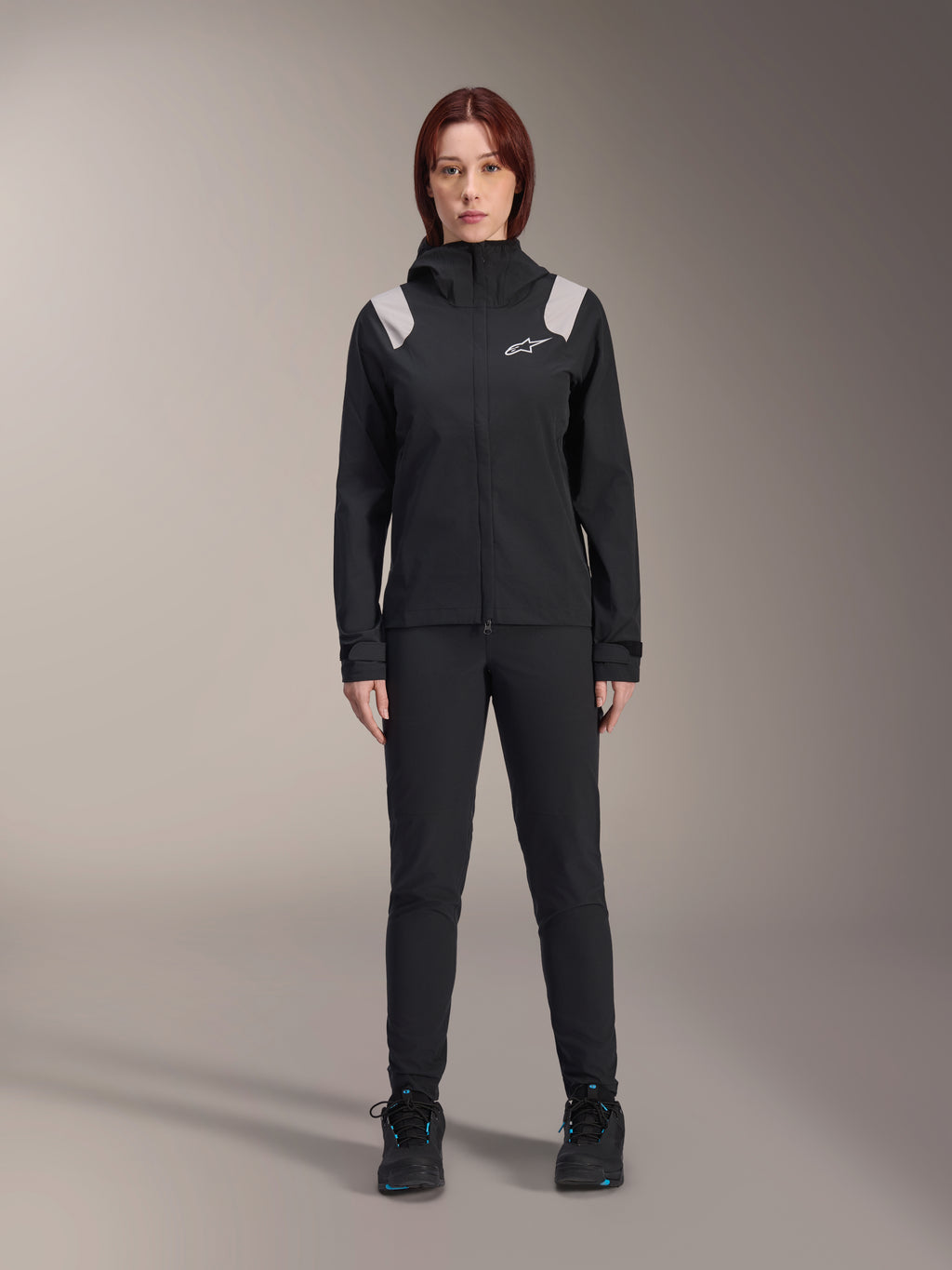 Alpinestars Stella A-Dura Rain Jacket, casaco de chuva de BTT para mulher, preto com detalhes cinzento claro nos ombros e logótipo branco, usado com calças e sapatos pretos a condizer, com capuz e punhos ajustáveis, concebido para a prática de BTT