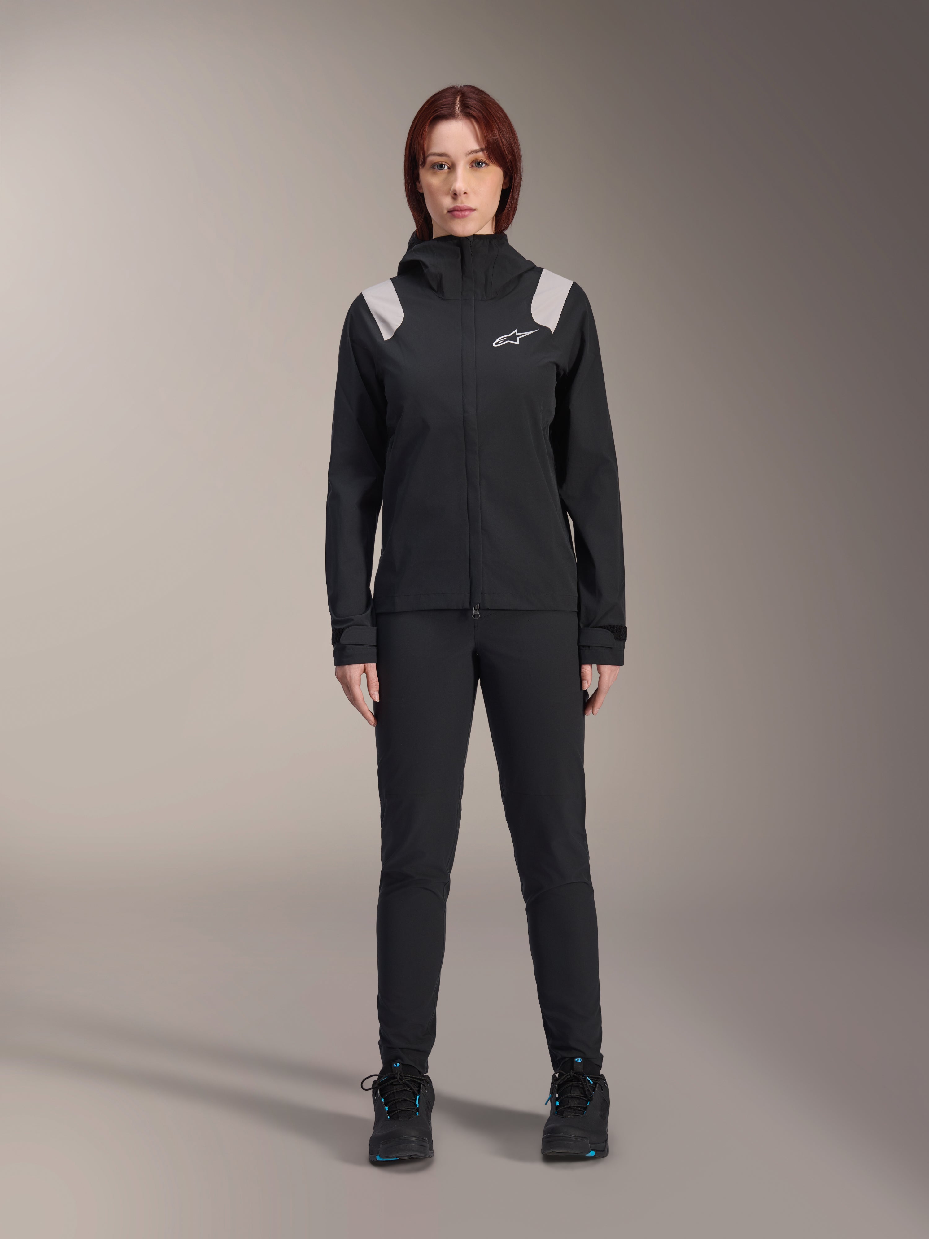 Alpinestars Stella A-Dura Rain Jacket, casaco de chuva de BTT para mulher, preto com detalhes cinzento claro nos ombros e logótipo branco, usado com calças e sapatos pretos a condizer, com capuz e punhos ajustáveis, concebido para a prática de BTT