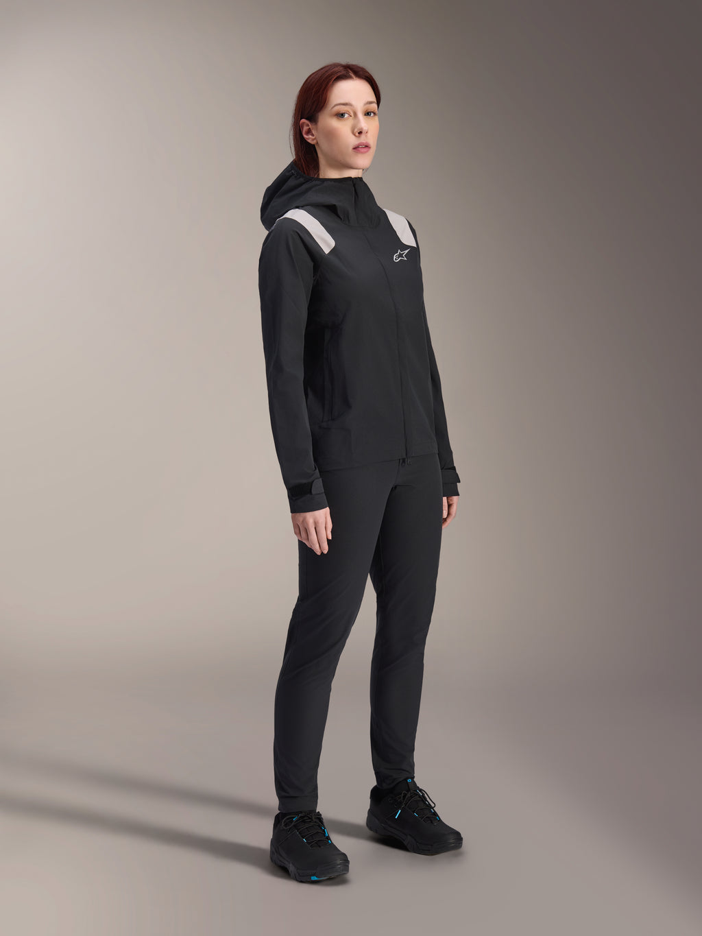 Mulher a usar o Casaco de Chuva Alpinestars Stella A-Dura em preto, casaco impermeável de 2,5 camadas para BTT com detalhes cinzento-claro nos ombros, calças pretas e sapatos de ciclismo a condizer, retrato de estúdio de equipamento de ciclismo de performance