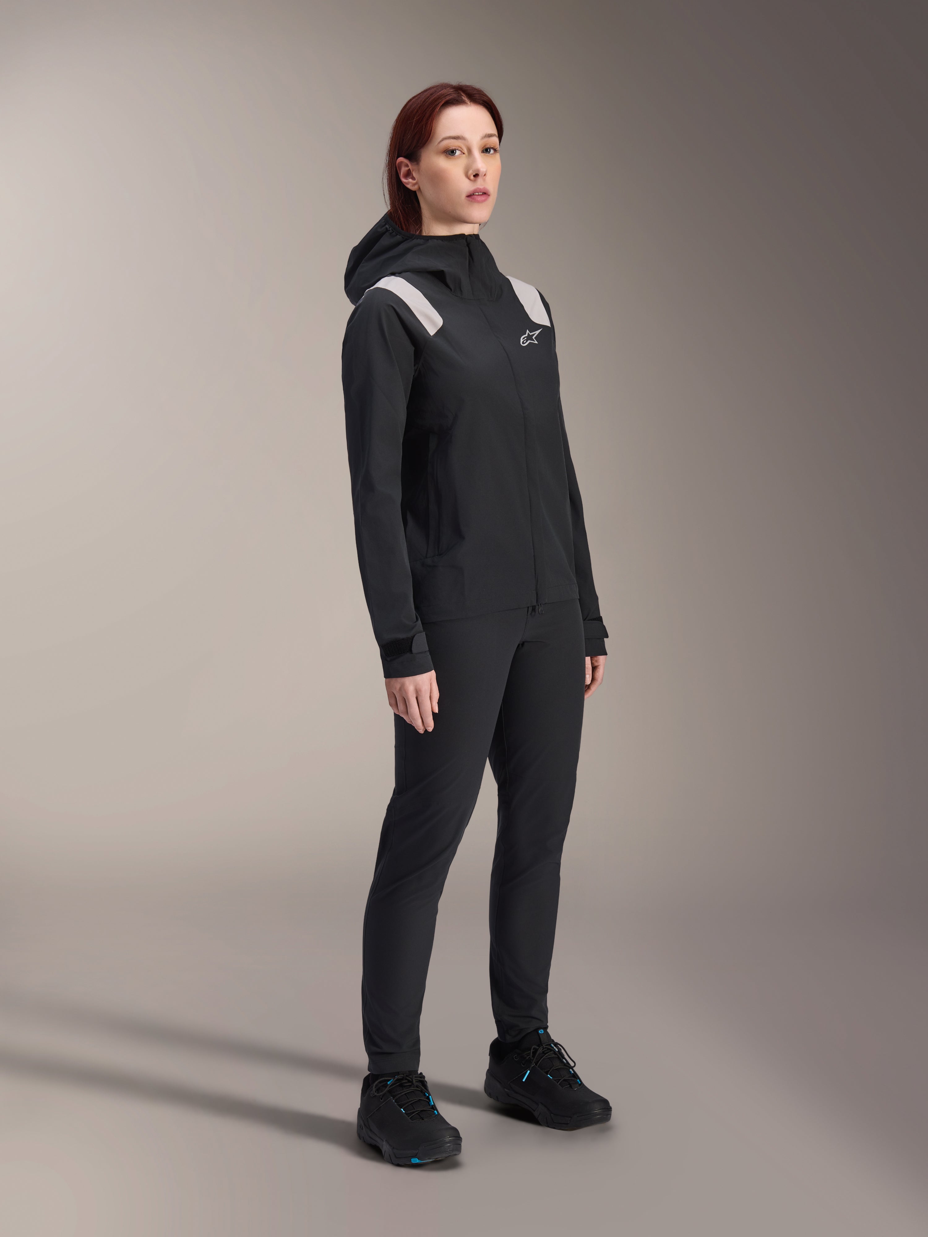 Mulher a usar o Casaco de Chuva Alpinestars Stella A-Dura em preto, casaco impermeável de 2,5 camadas para BTT com detalhes cinzento-claro nos ombros, calças pretas e sapatos de ciclismo a condizer, retrato de estúdio de equipamento de ciclismo de performance