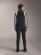 Ciclista vista de costas a usar o Casaco de Chuva Alpinestars Stella A-Dura, preto com painéis cinzento claro nas mangas e ombros, capuz levantado, combinado com calças pretas e sapatos de ciclismo, equipamento técnico de chuva para BTT